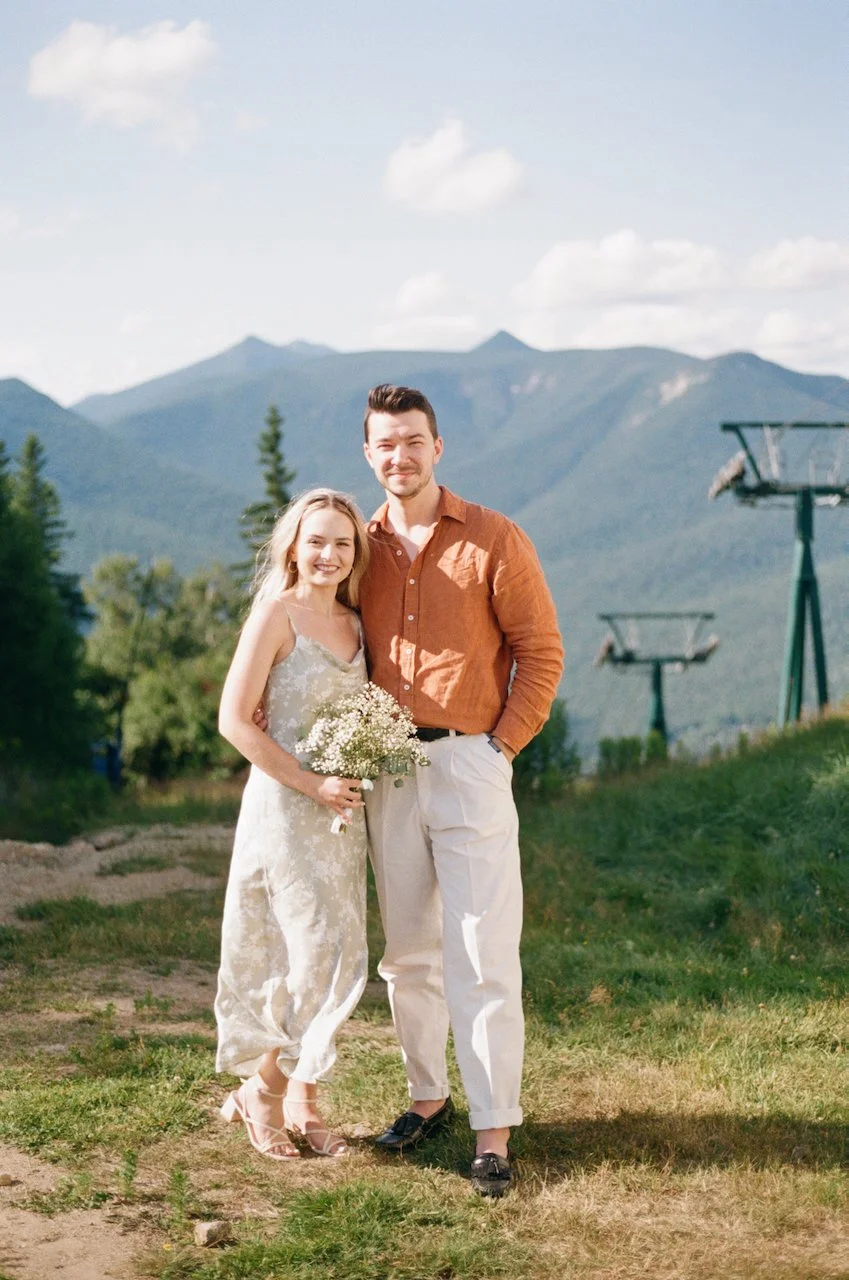 hayden budd photo + film A smiling couple standing outdoors on a grassy area with mountains in the background. The woman is wearing a white dress and holding a small bouquet of white flowers, while the man is dressed in a brown shirt and light pants.