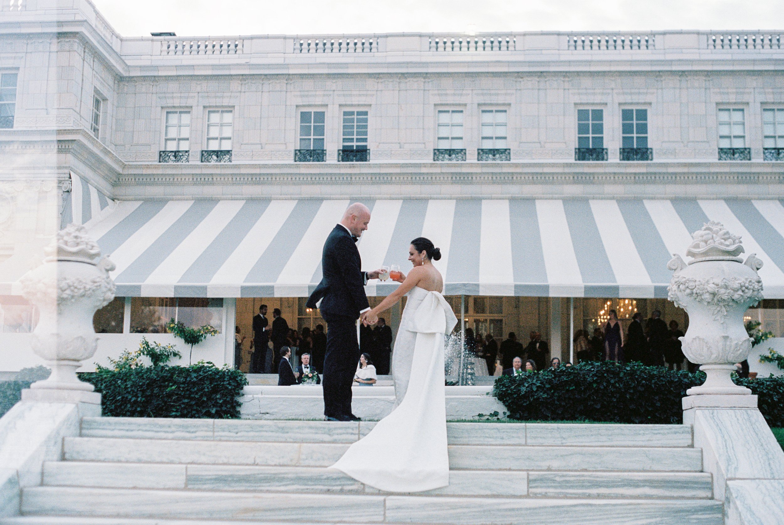 A bride and groom holding hands and exchanging glasses of champagne on the steps of a luxurious building during their wedding celebration.