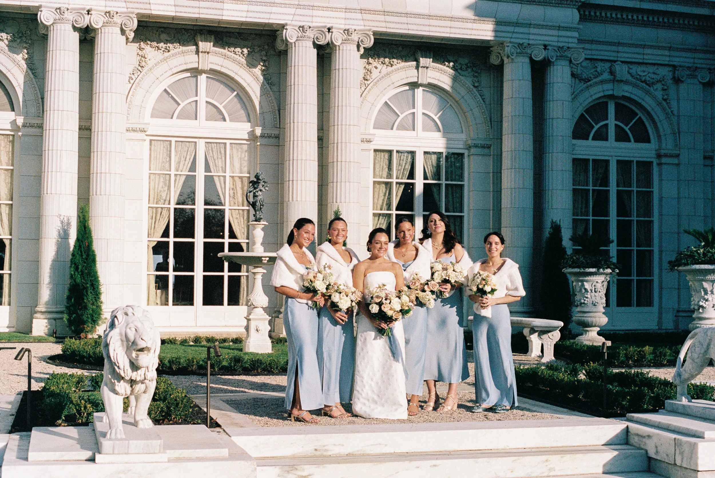 A bride in a strapless white wedding gown stands with five bridesmaids in pale blue dresses, all holding bouquets of white and blush roses, in front of a grand, ornate mansion hayden budd photo + film new england film photographer