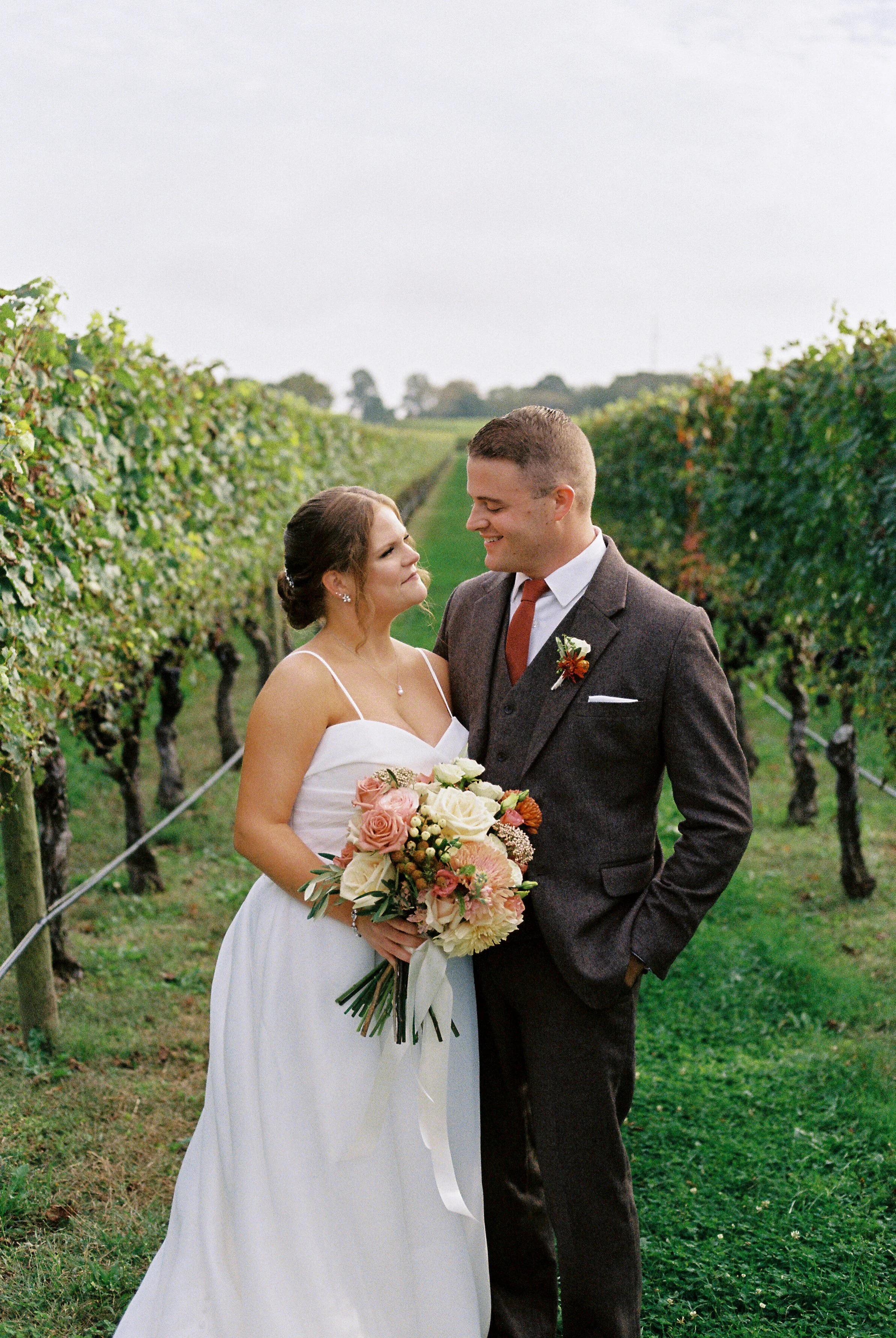 A bride and groom standing in a vineyard, gazing at each other. The bride is holding a bouquet of pink and white flowers, and the groom is smiling. The vineyard has rows of grapevines on either side. hayden budd photo + film new england film photogra