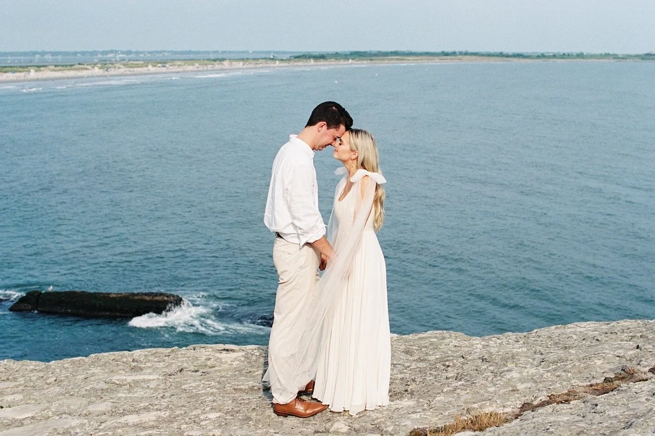 hayden budd photo + film new england film photographer A couple dressed in white standing close on a rocky coastline by the water, with a distant shoreline and no other people visible.
