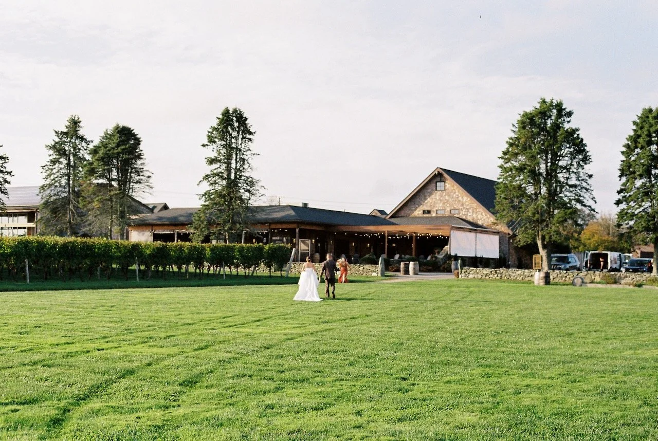 A bride and groom walking on a grassy field towards a rustic building with string lights, with a few guests nearby and parked cars in the background. hayden budd photo + film new england film photographer