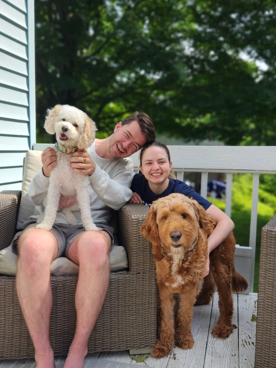 hayden budd photo + film new england film photographer A happy couple with two dogs sitting on a porch. The man is holding a white poodle, and the woman is leaning on a golden doodle. The porch has wicker furniture and is surrounded by green trees.