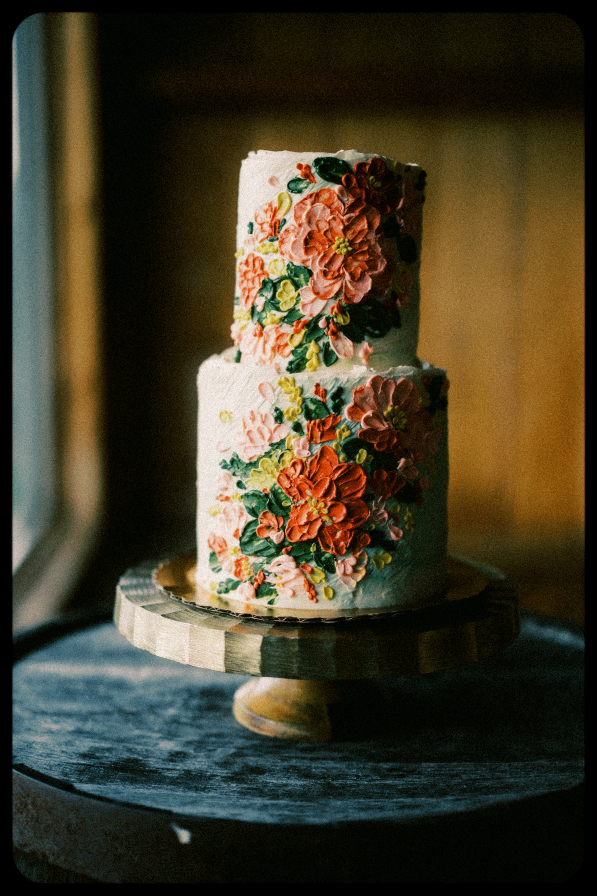 A two-tiered wedding cake decorated with colorful flower icing designs, placed on a wooden cake stand near a window. hayden budd photo + film new england film photographer