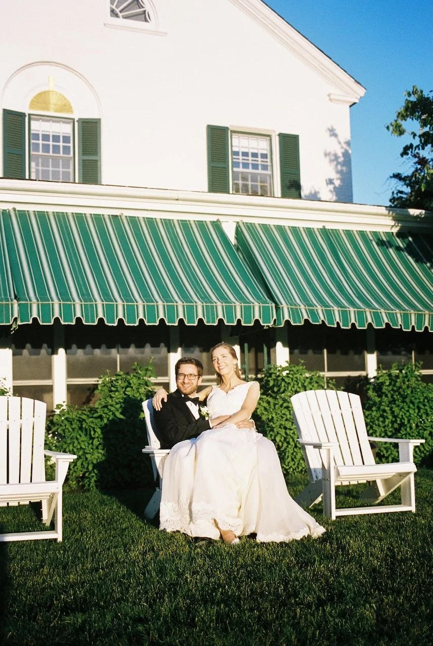 A newlywed couple sitting on a white bench outdoors in front of a building with green shutters and a green-striped awning, smiling and enjoying the sunny day. hayden budd photo + film new england film photographer