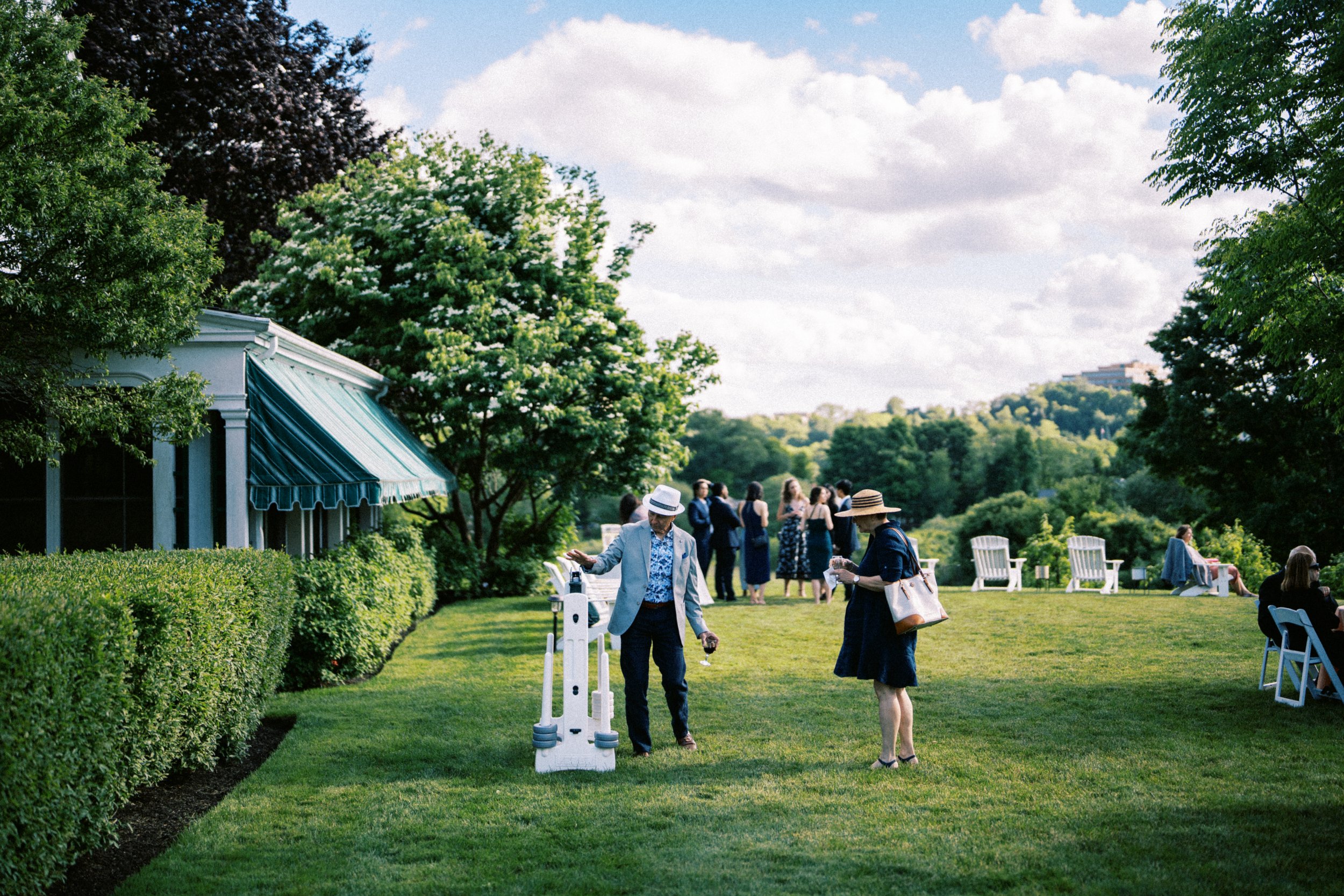 People attending an outdoor social gathering in a lush green park with trees, chairs, and a building with a blue awning. hayden budd photo + film new england film photographer