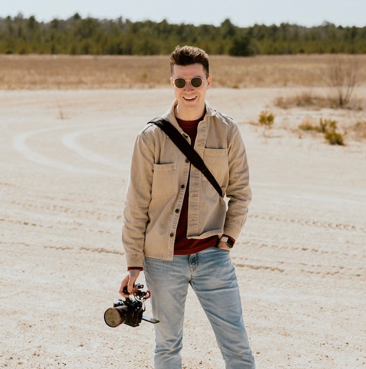 hayden budd photo + film new england film photographer  standing in a desert landscape holding a camera, wearing sunglasses, a beige jacket, and jeans