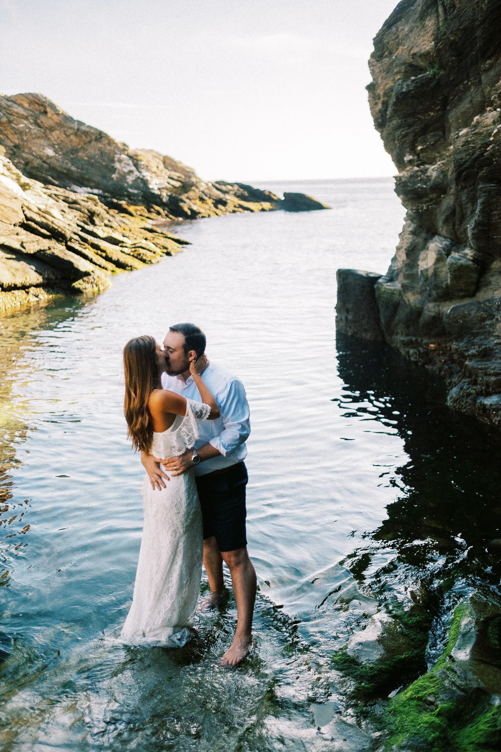 A couple sharing a kiss in a rocky coastal area with water and cliffs in the background.