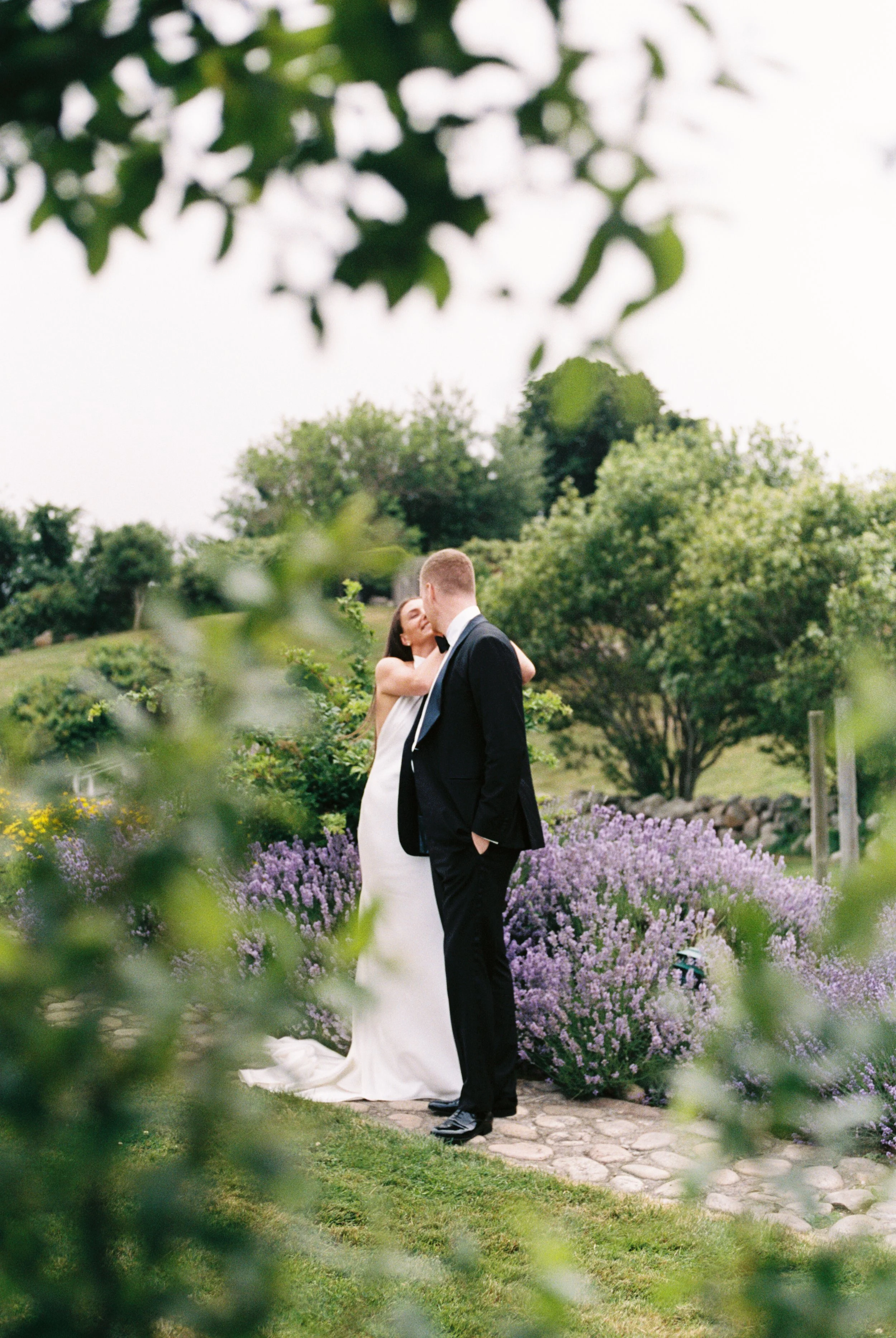 A bride and groom kiss outdoors, framed by greenery, on a cobblestone path surrounded by purple flowers and trees.
