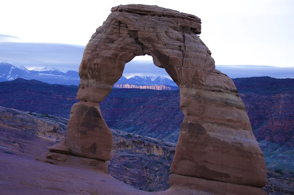Arches National Park "Delicate Arch" Sunrise with the sun glowing off the far red rocks beneath the snow capped La Sal mountains.