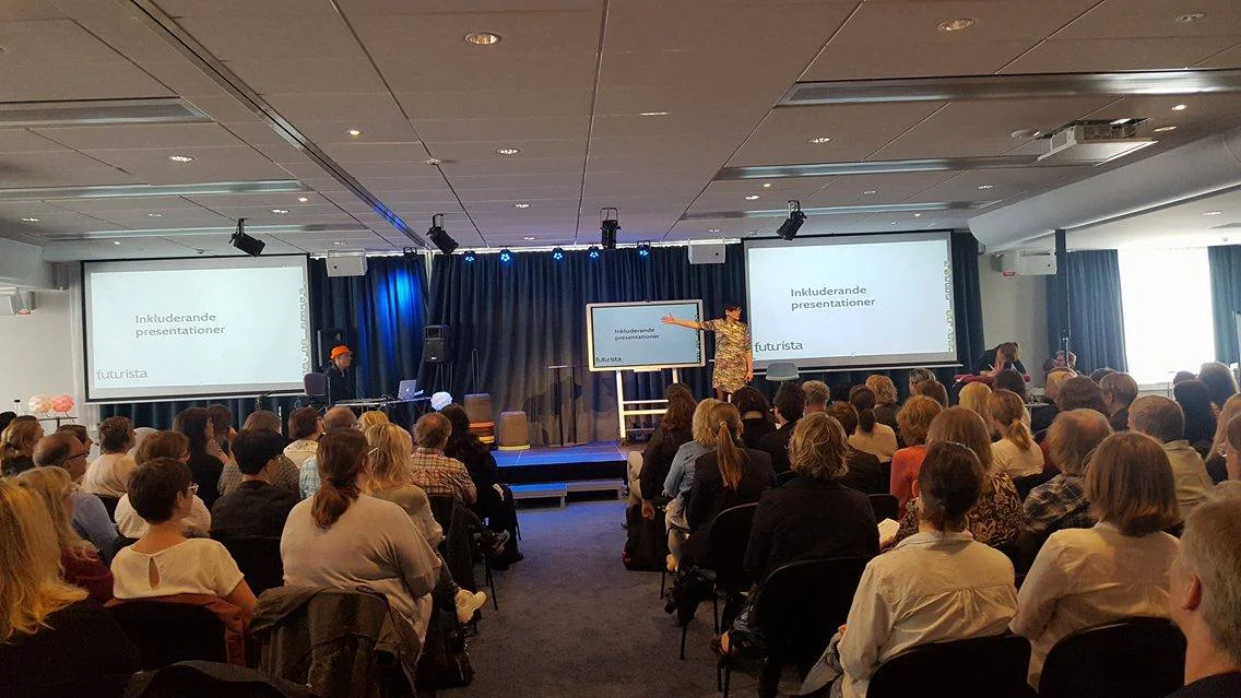 Publik sedd bakifrån i förgrunden. Åsa på scenen med en arm rakt utsträkt åt sidan. På skärmarna står det Inkluderande presentationer