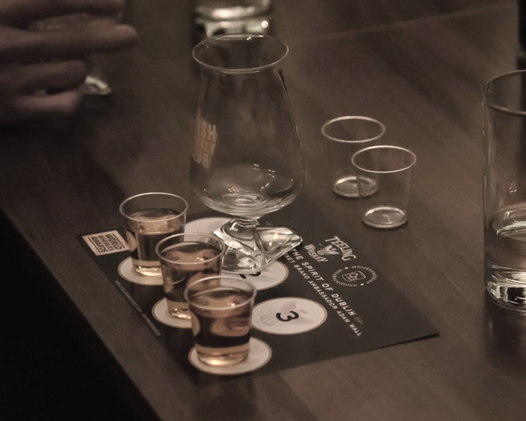 A dark table with a glass water pitcher, multiple shot glasses filled with a dark liquid, and a glass of water. The table has printed text and circles with numbers, possibly for a whiskey tasting or similar event.
