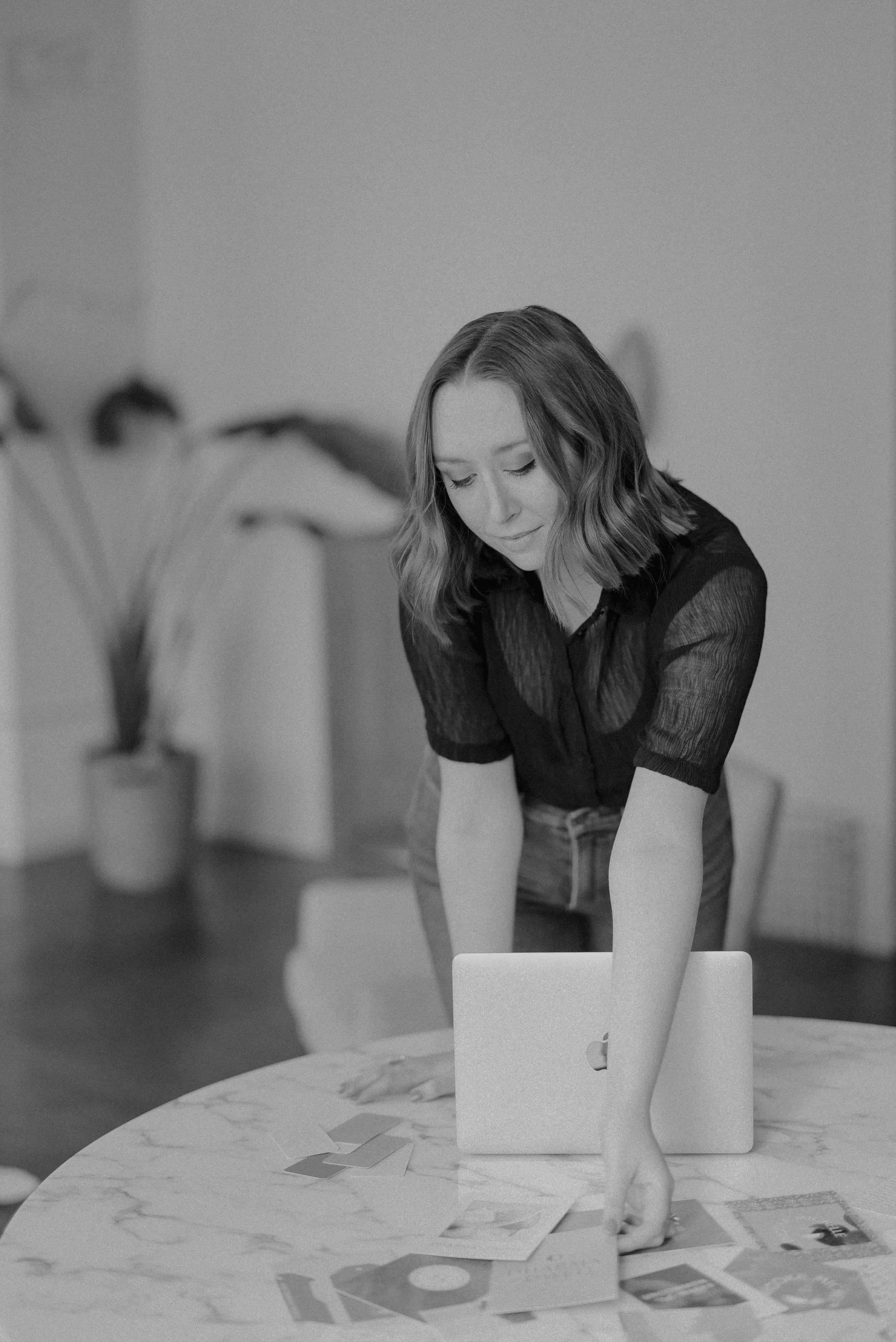 A woman with shoulder-length wavy hair and wearing a dark short-sleeved shirt is looking down, working on a laptop placed on a round marble table. There are papers and photos scattered on the table. The background shows a blurred figure in front of a light-colored wall with two houseplants.