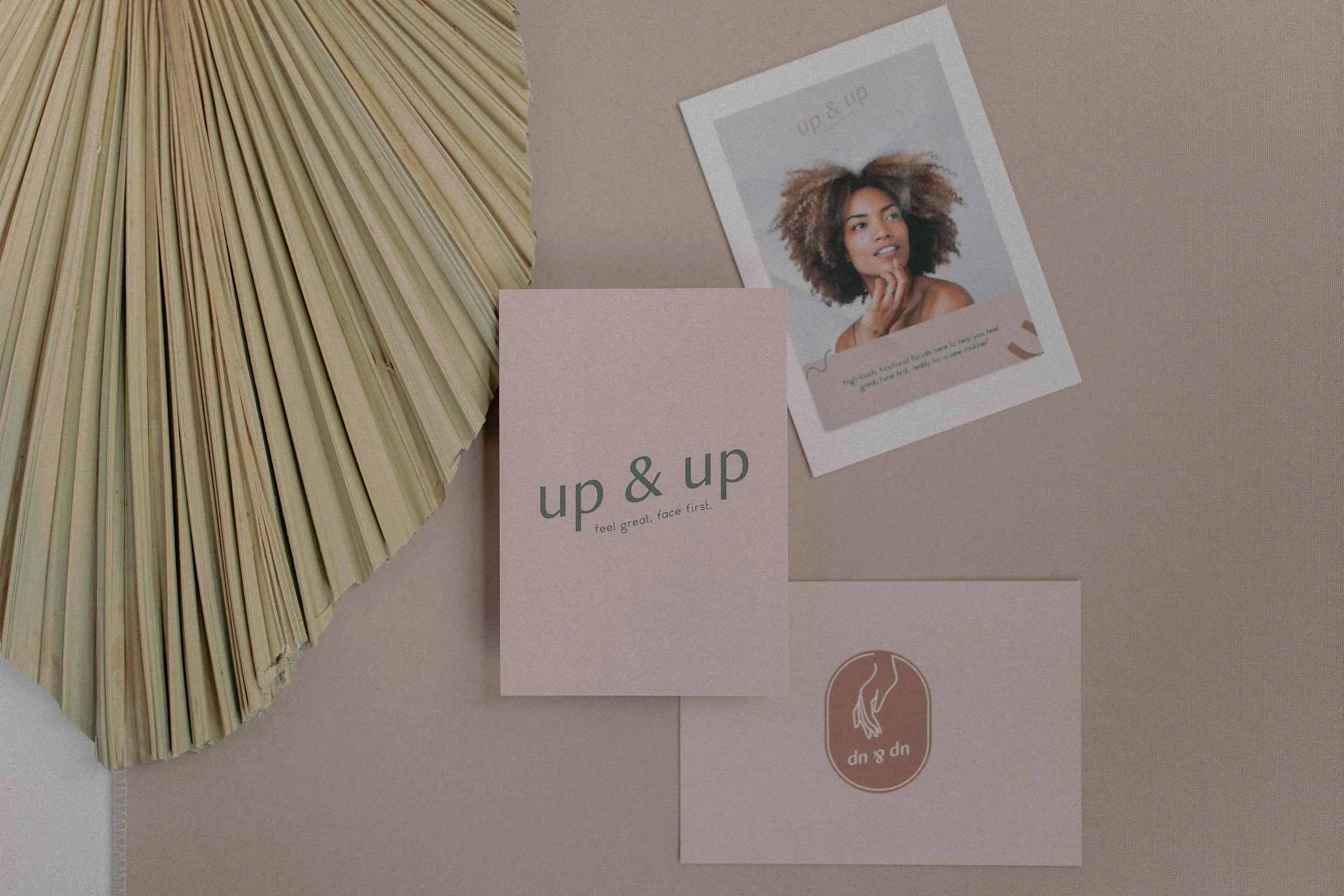 A fan with light-colored blades, a brochure titled 'up & up' with the subtitle 'feel great, face first,' and a flyer with 'up & up' printed on it along with an image of a woman with curly hair touching her chin.