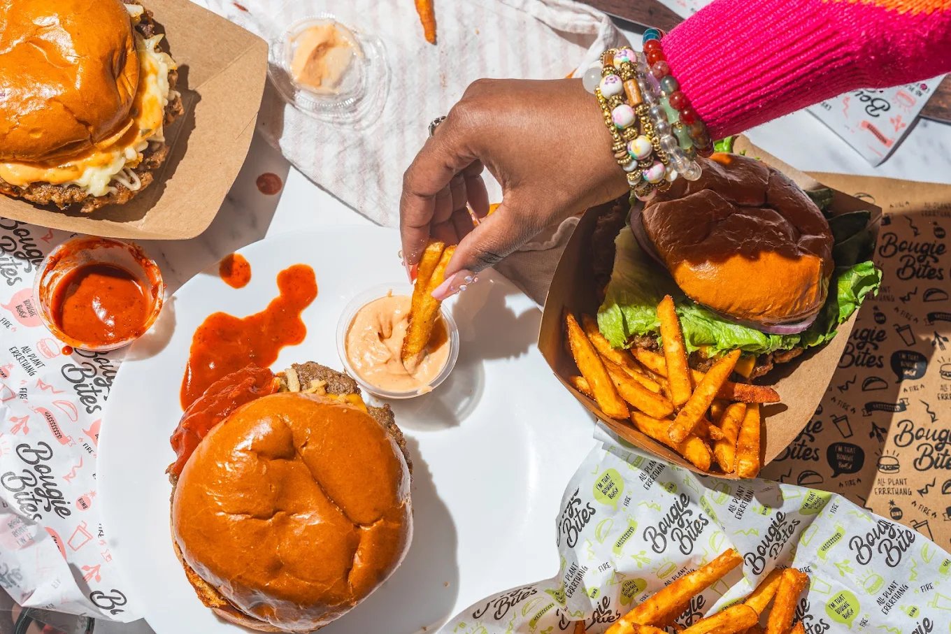 A table with two burgers, fries, ketchup, and a woman dipping a fry into a spicy orange sauce.