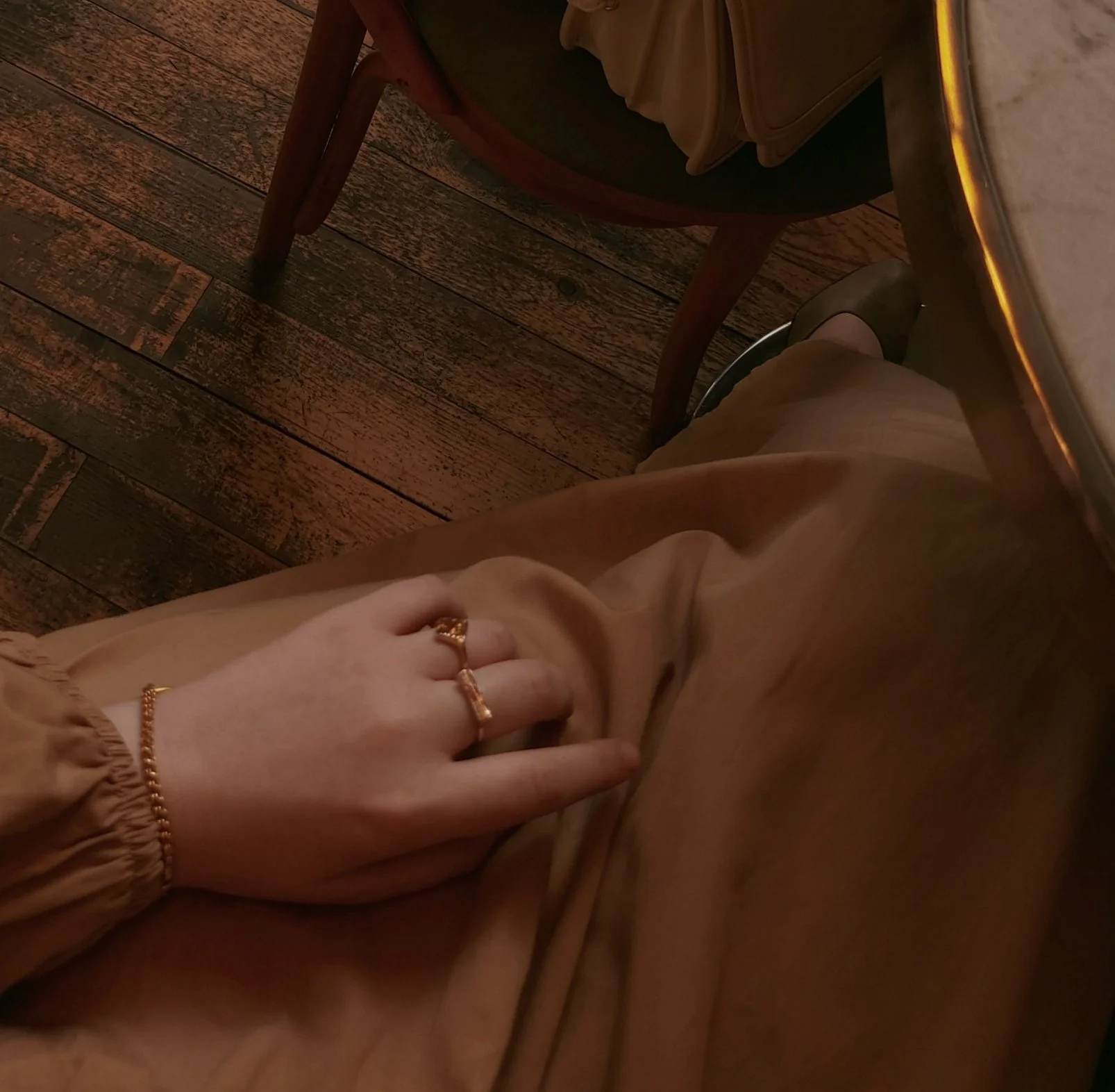 A person wearing beige pants with gathered cuffs, sitting at a wooden table. Their left hand, adorned with a gold ring and bracelet, rests on their thigh. The table has a yellow edge trim, and a chair with a wooden frame and dark green cushion is visible. The background features a wooden floor.