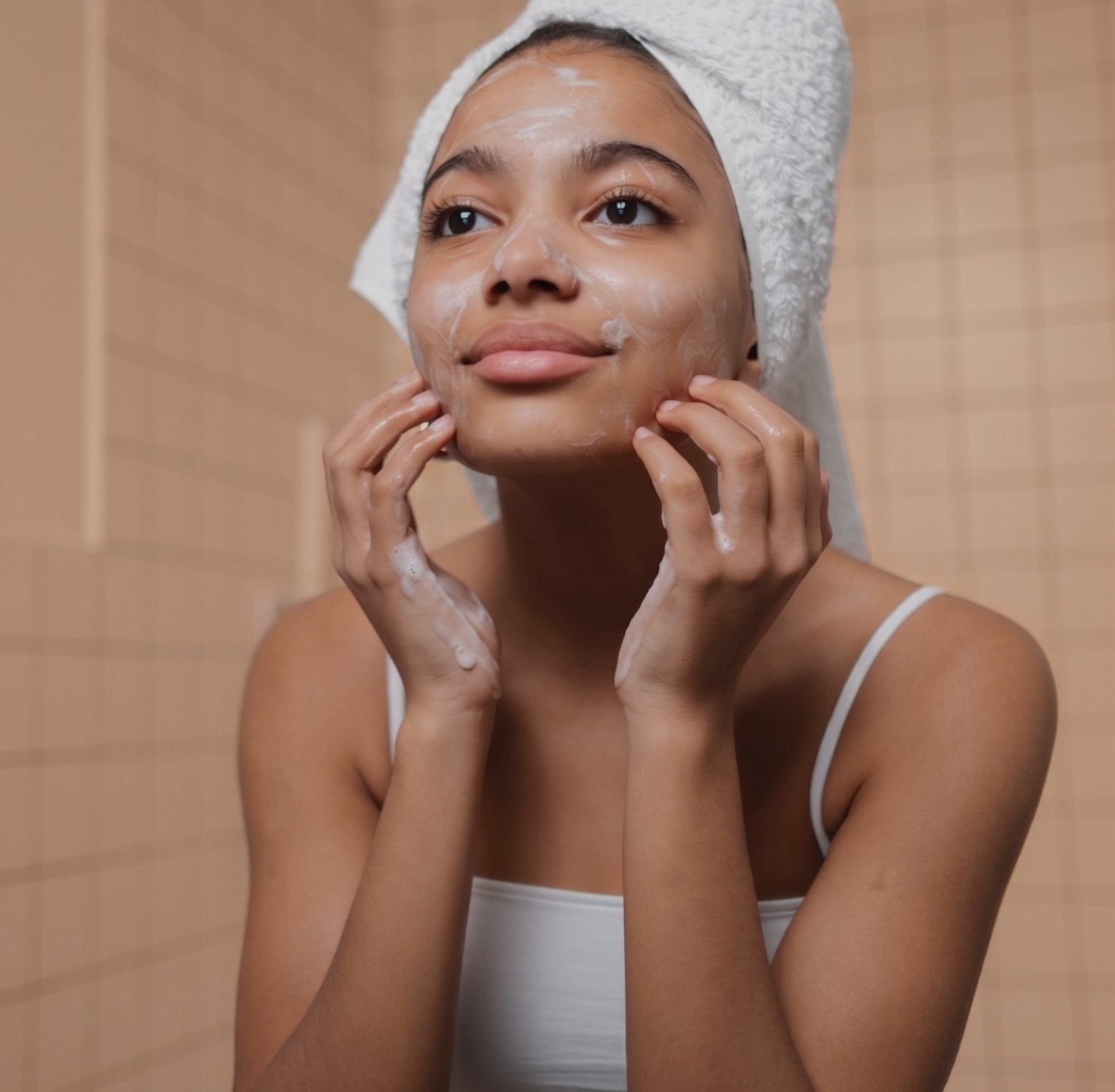 A woman with a towel wrapped around her head is washing her face in a shower or bathroom with beige tiled walls.