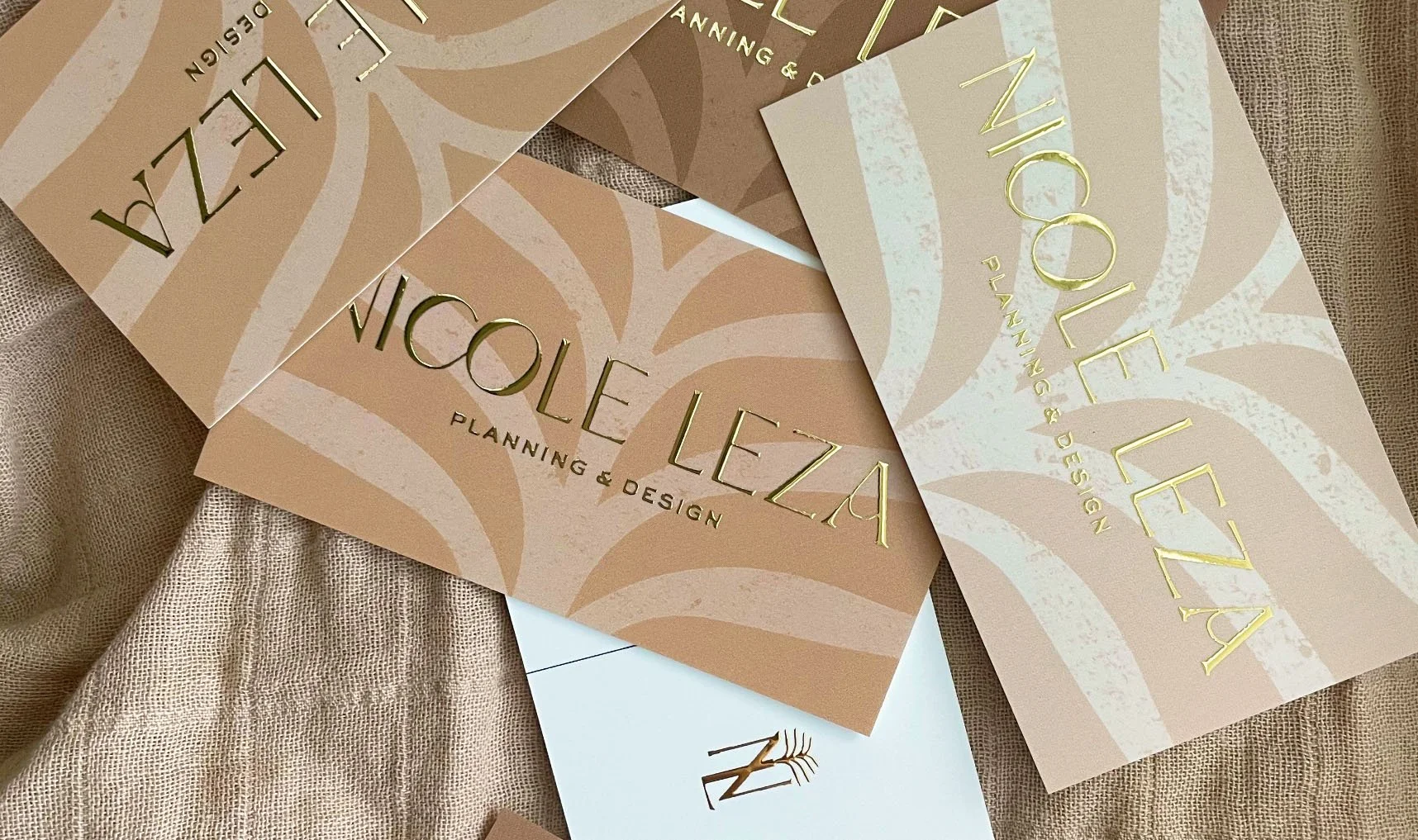 Business cards for Nicole Leza Planning & Design with a beige and gold color scheme, laid on textured beige fabric.