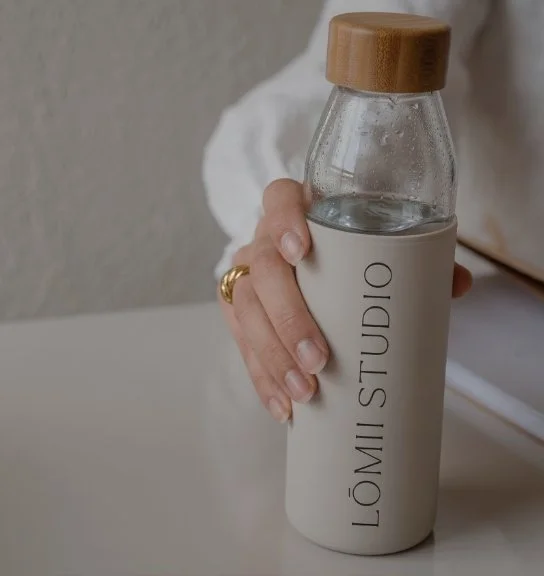 A person holding a glass water bottle with a wooden lid and a white sleeve labeled 'LOMI STUDIO'.