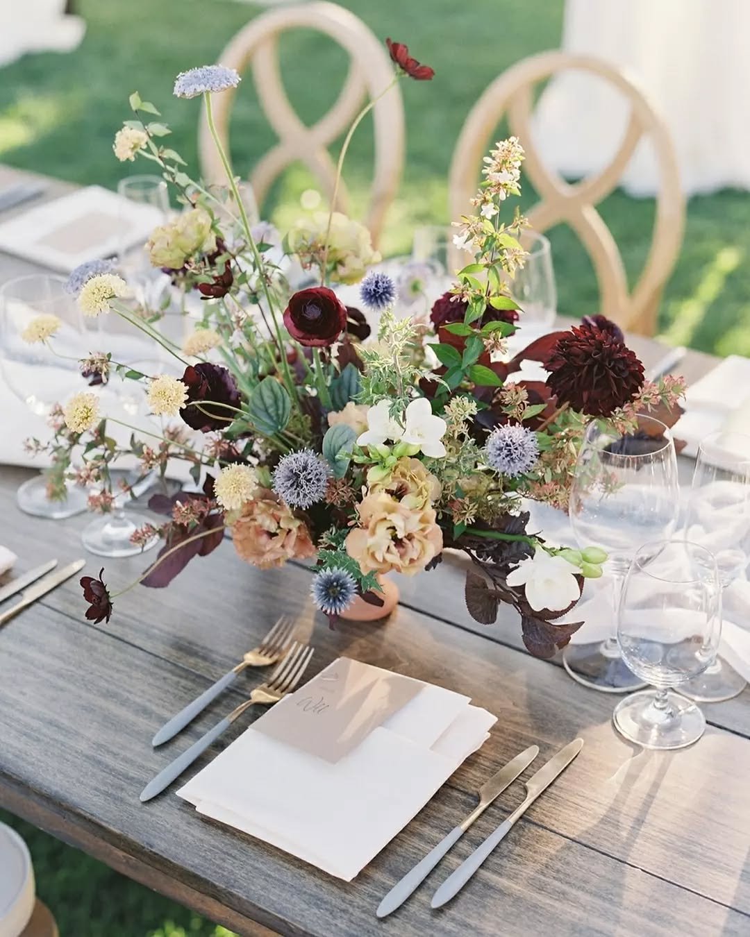 Alfresco dining never looked so inviting. 🌿 These stunning floral centerpieces, with rich jewel-toned burgundy, soft periwinkle, and creamy accents, brought effortless elegance to rustic wooden tabletops. 

A perfect balance of color and texture for