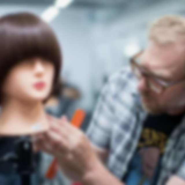 A stylist is applying makeup or styling to a mannequin head with brown hair, while a man with glasses looks on.
