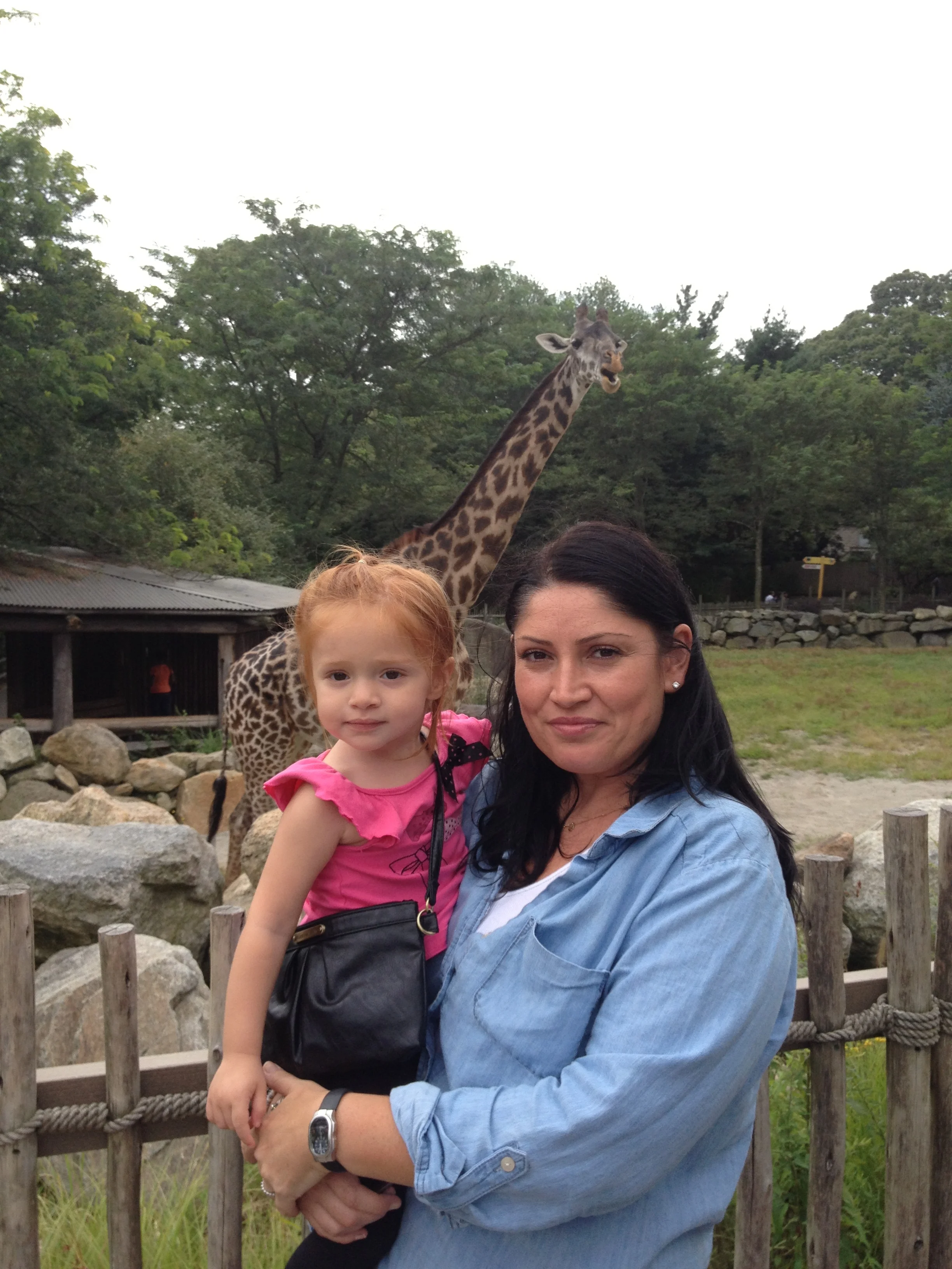 A woman holding a young girl in front of a giraffe at a zoo or wildlife park, with trees and rocks in the background.