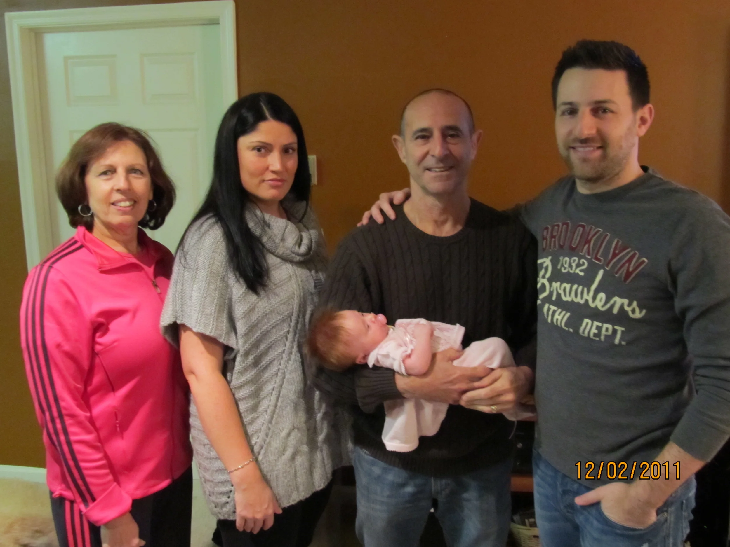 Five adults and a baby pose together indoors, with a woman in a pink athletic jacket on the left, a woman in a gray sweater next, a man holding a newborn dressed in pink, and a man in a gray sweatshirt on the right.