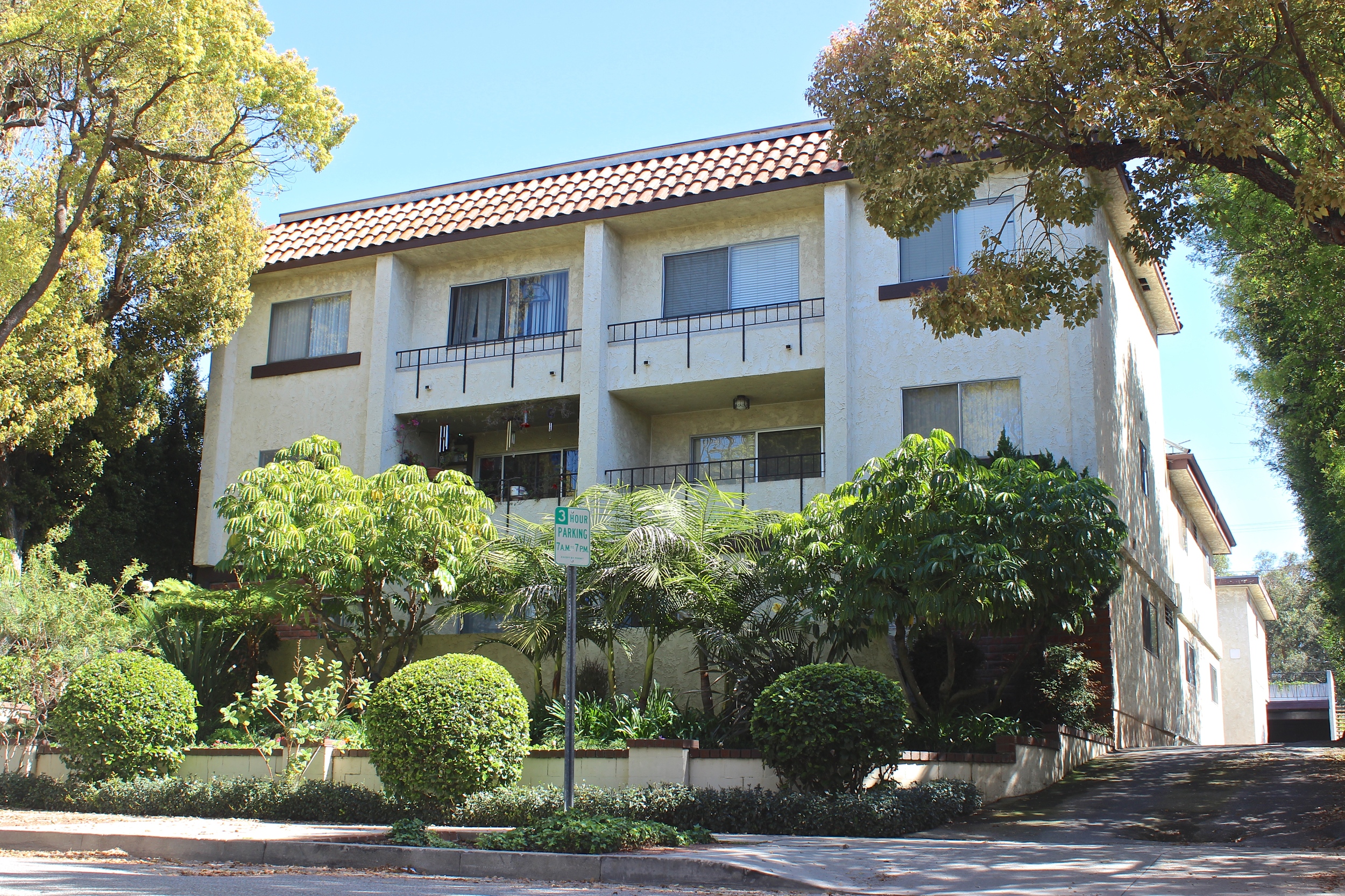 Street view of trees and shrubs surrounding three story off white with driveway leading to rear of the building.