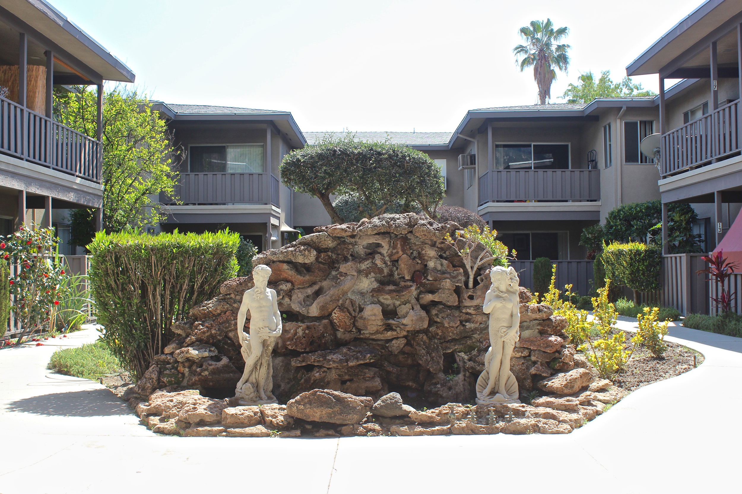 Stone water feature with white statues surrounded by two story gray apartment complex.
