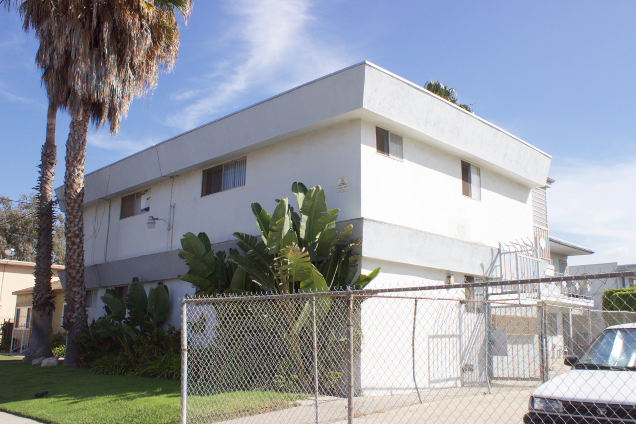 Stark white two-story building with palm tree and chain-link fence.