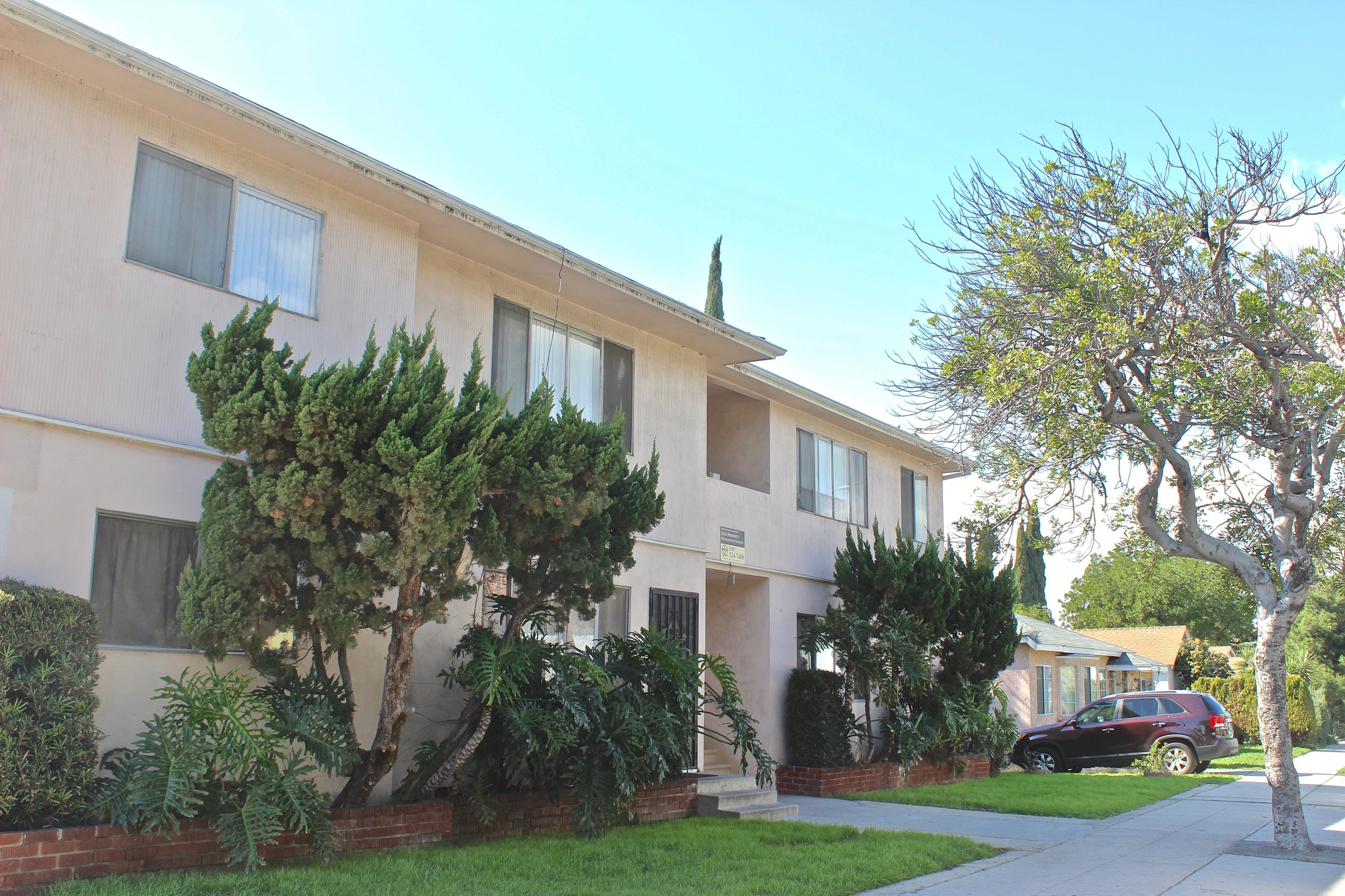  Two story off white apartment building  with trees and a sidewalk in front.