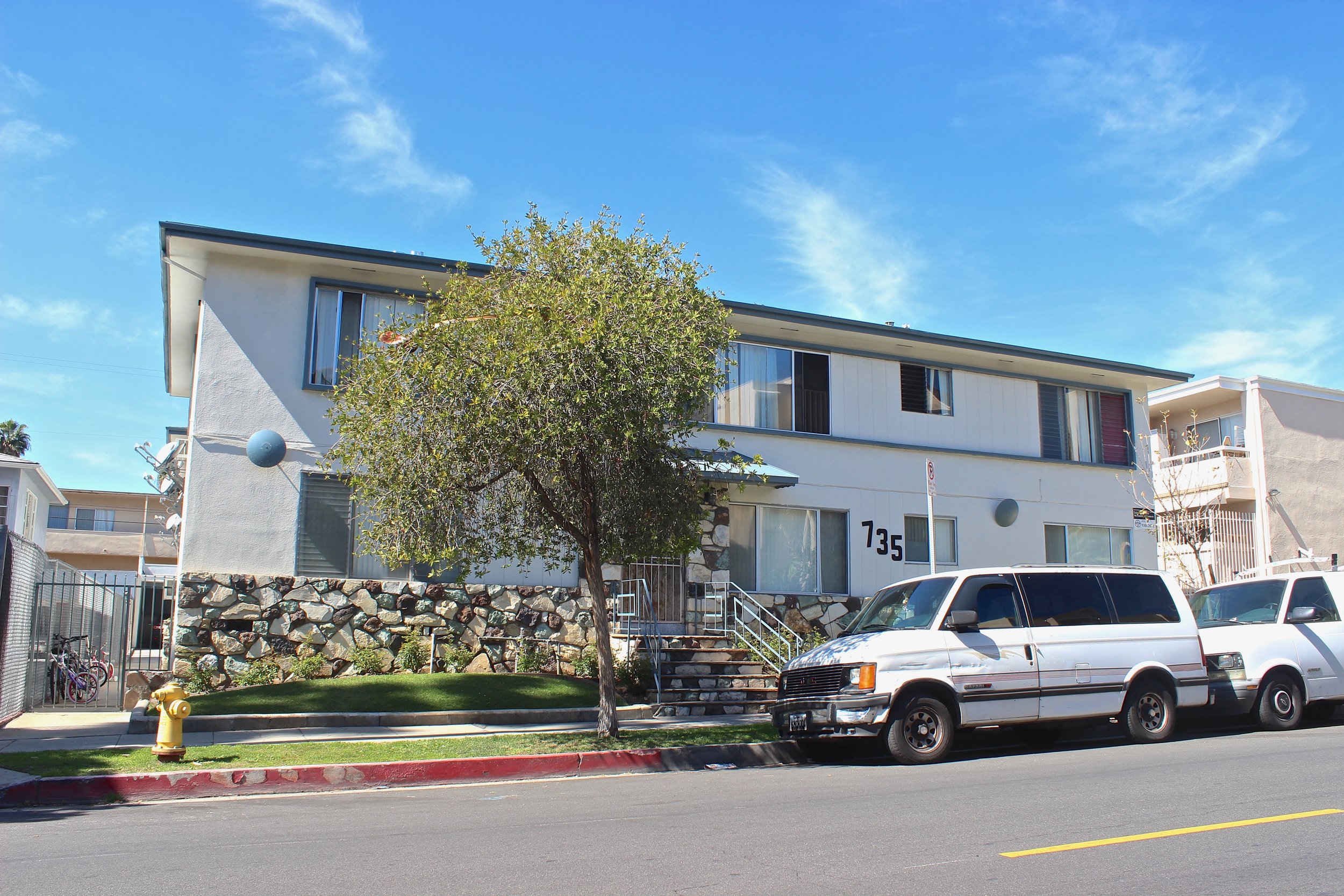 Two story white apartment building with green accents and stone facade on the first floor with street parking. 