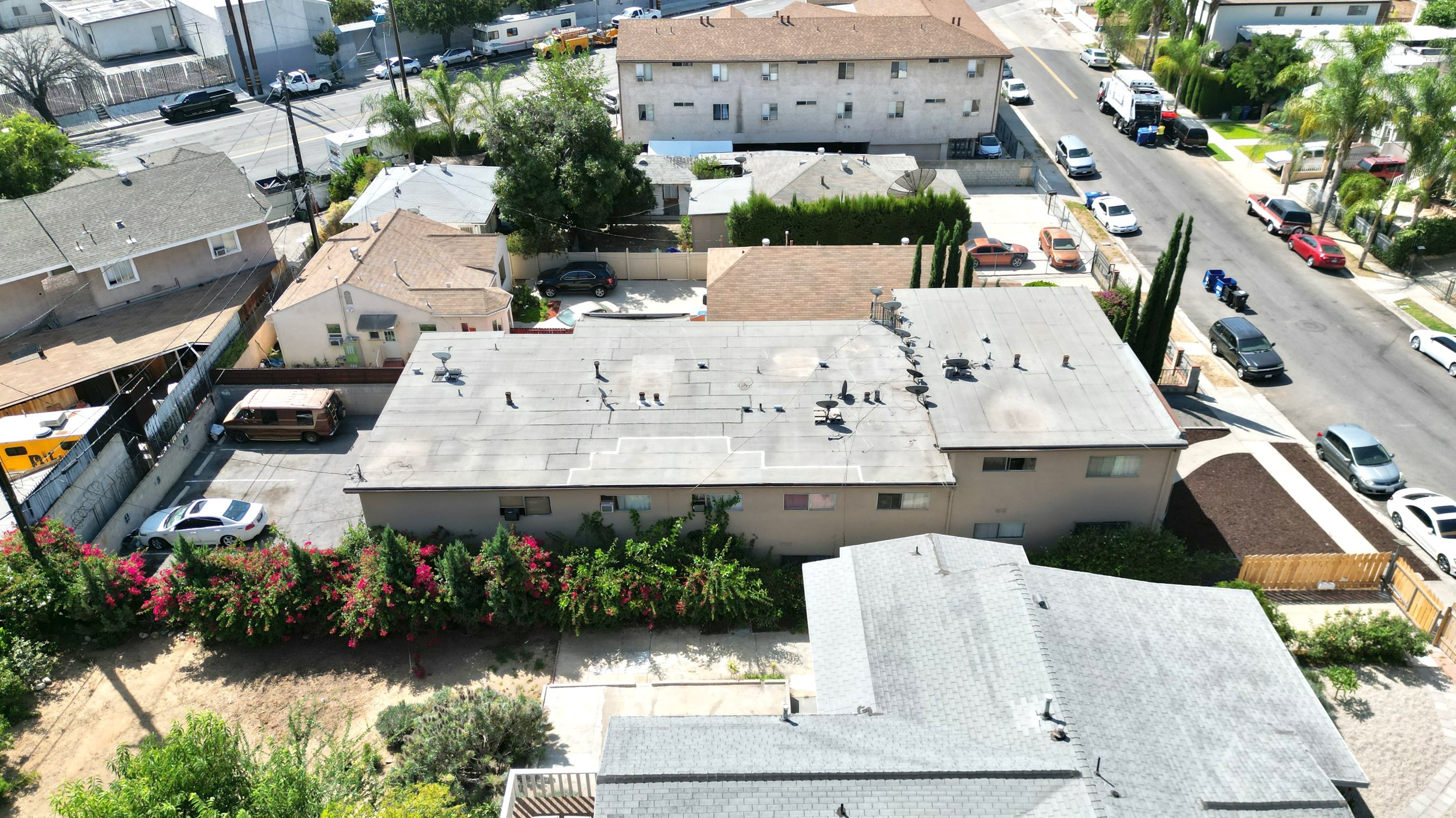 Aerial view of a residential block with buildings, flowering bushes, and street parking.