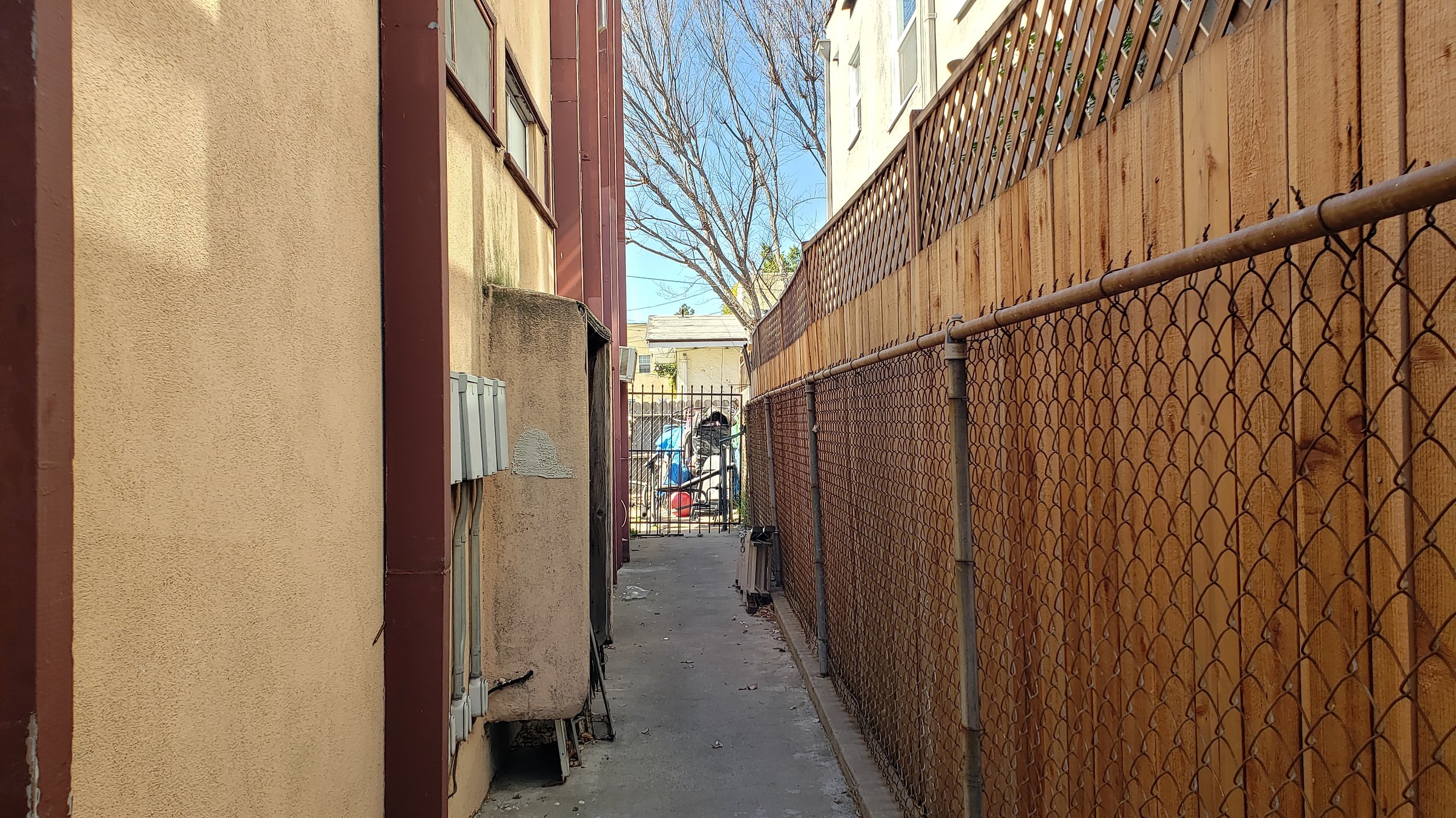 Narrow alley with chain-link fence and rear view of residential buildings.