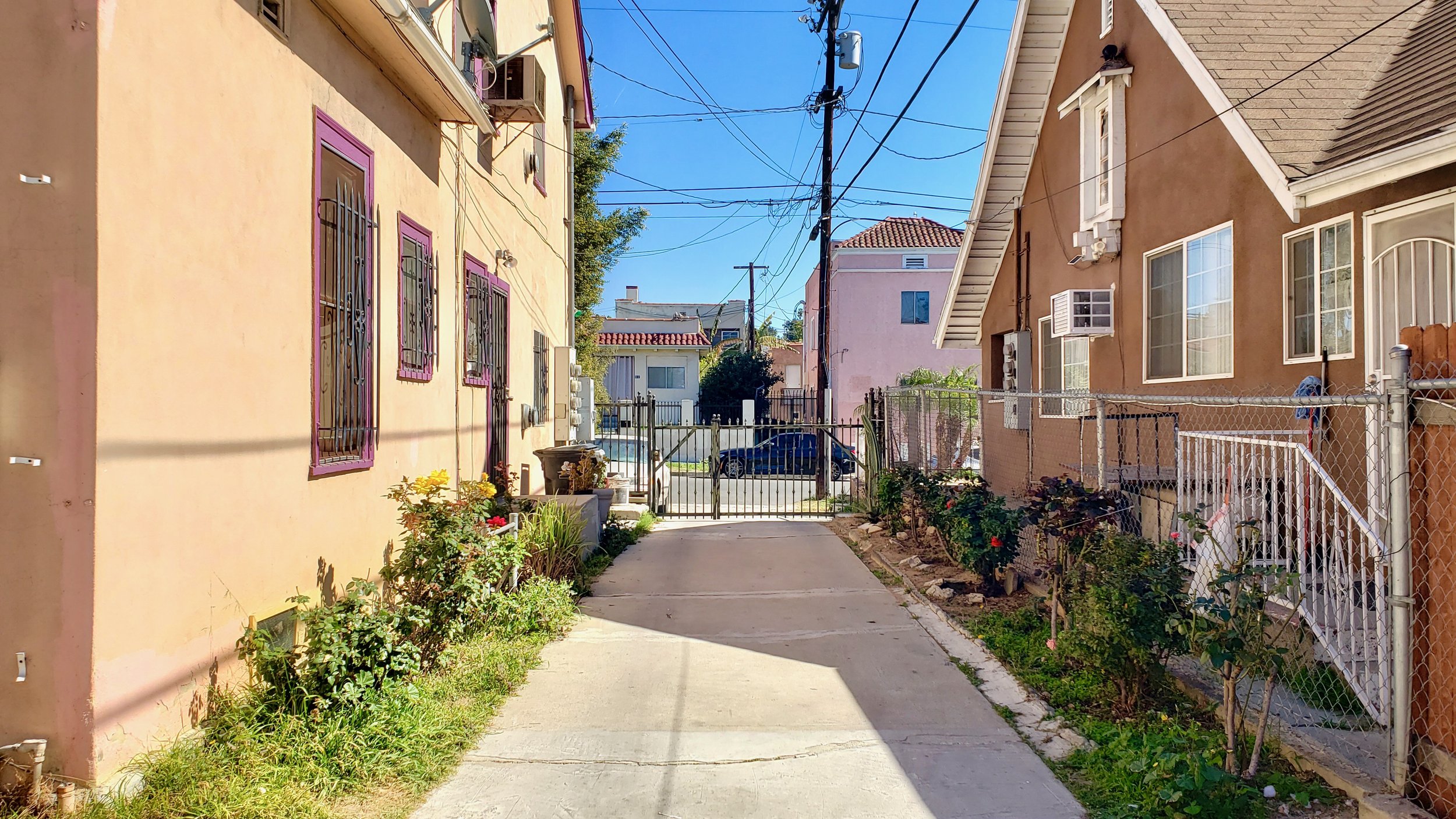 A residential alley with a pink apartment, gated path, and flower beds under a clear sky.
