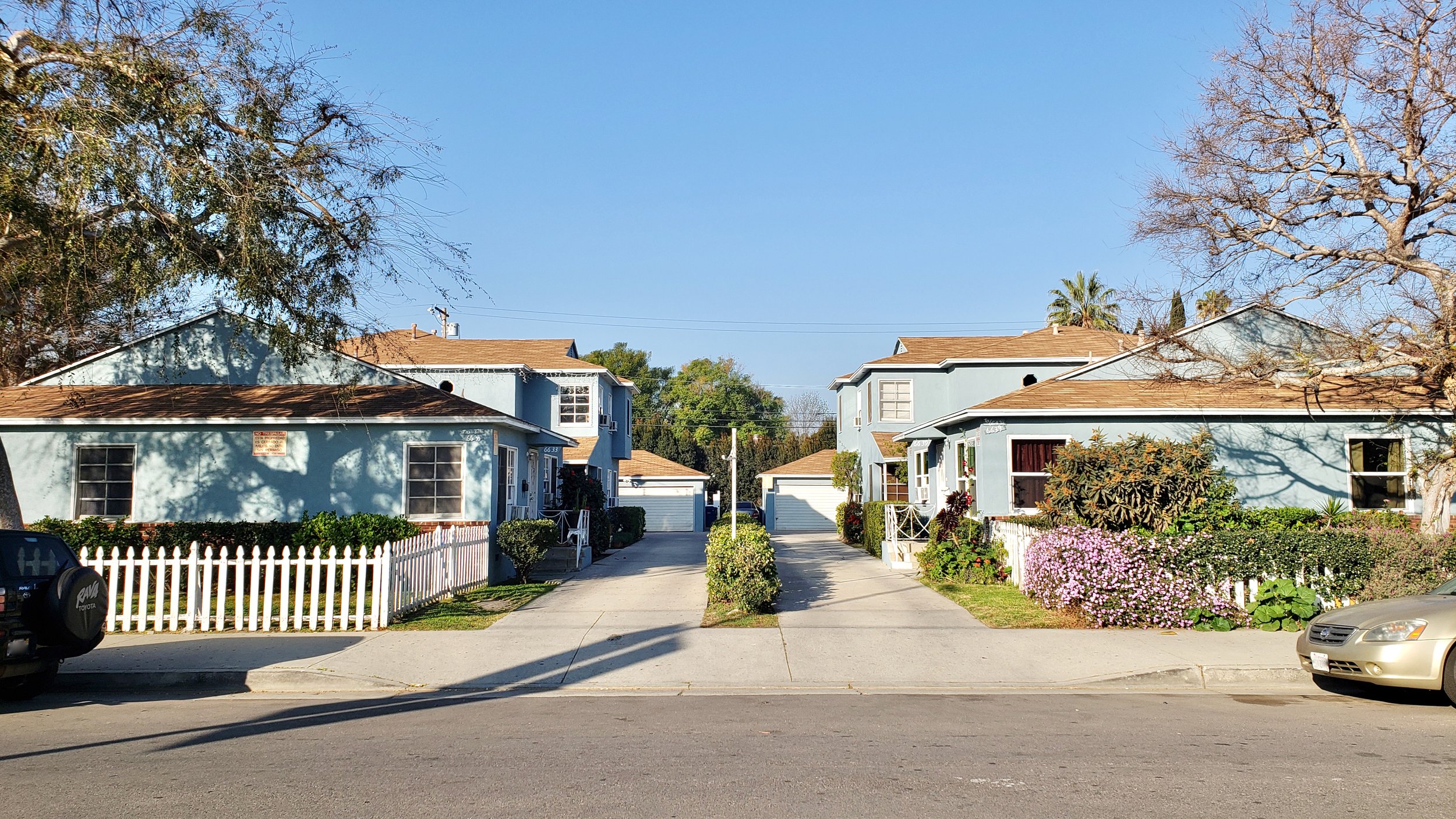 A sunny street view showing quaint houses with driveways and a white picket fence.