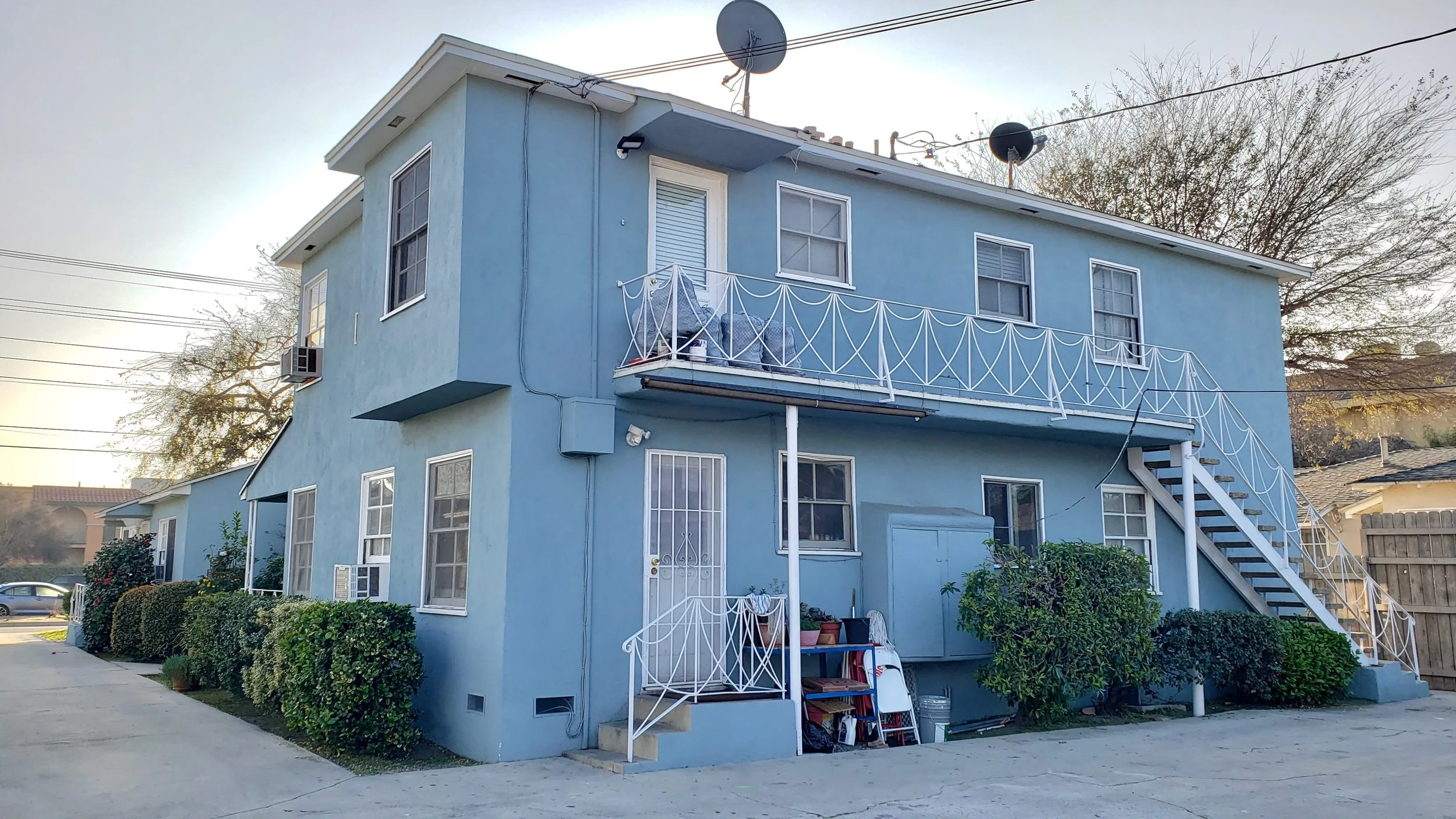 Rear view of a two-story light blue home with external stairs and a balcony, featuring lush green shrubbery and clear skies.