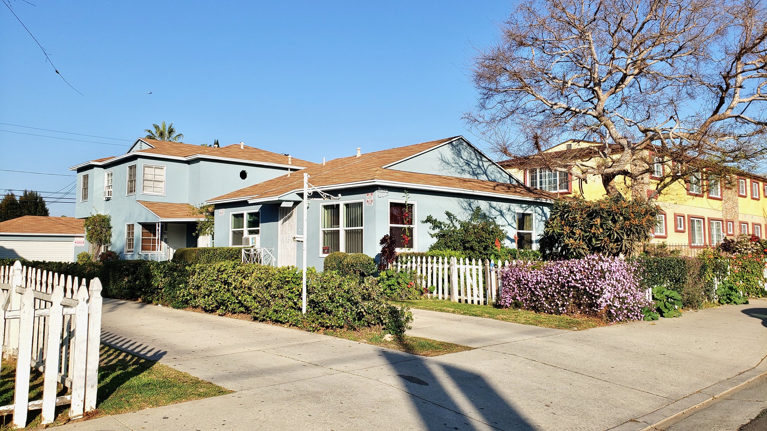 A serene residential scene showing a blue single-story house next to a two-story building under clear blue skies and trees.