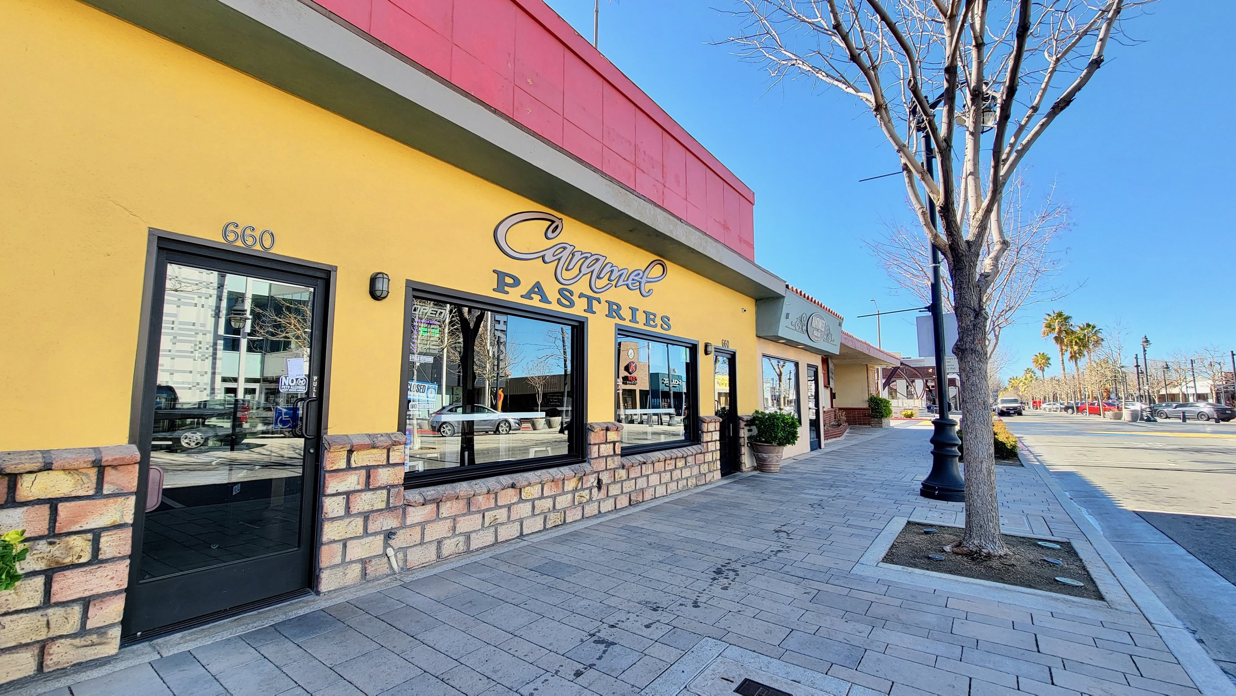 Sidewalk view of Caramel Pastries shop with yellow walls and a bright sky, leafless tree nearby.