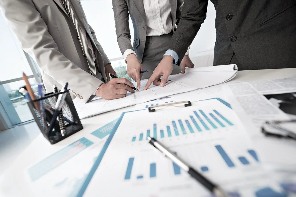 Professionals examining architectural plans on a desk with financial charts.
