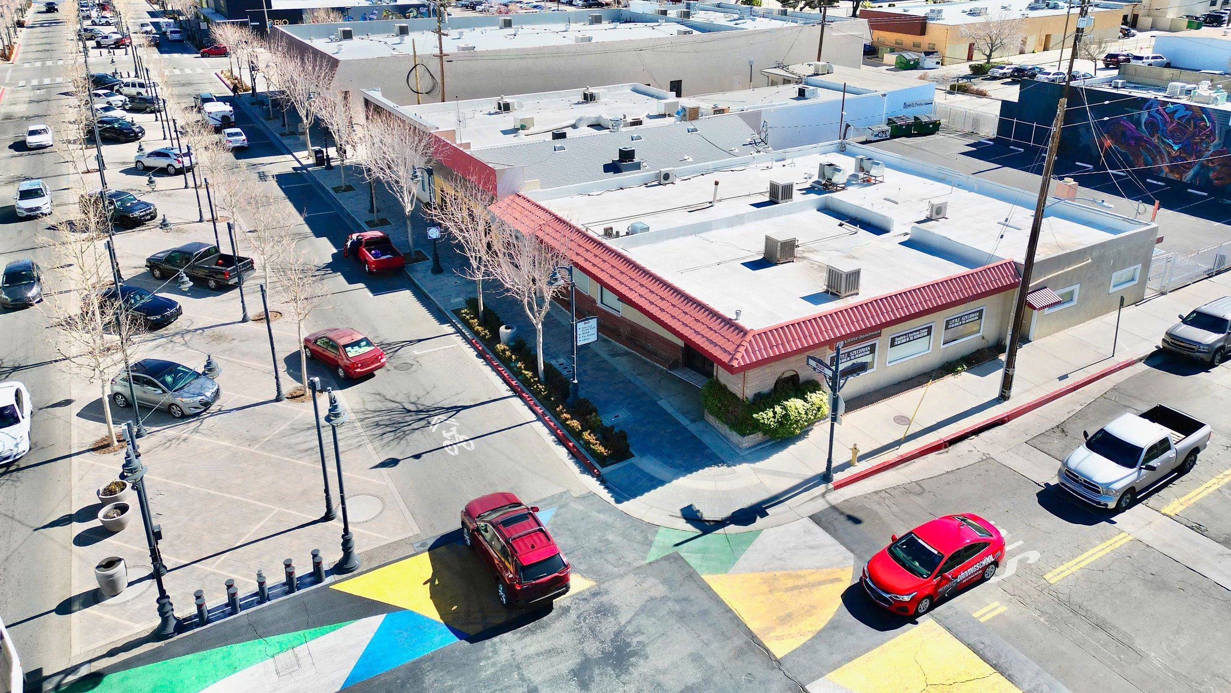 An aerial view of a street intersection with colorful street art, a red-roofed building, and parked cars, under a clear sky.