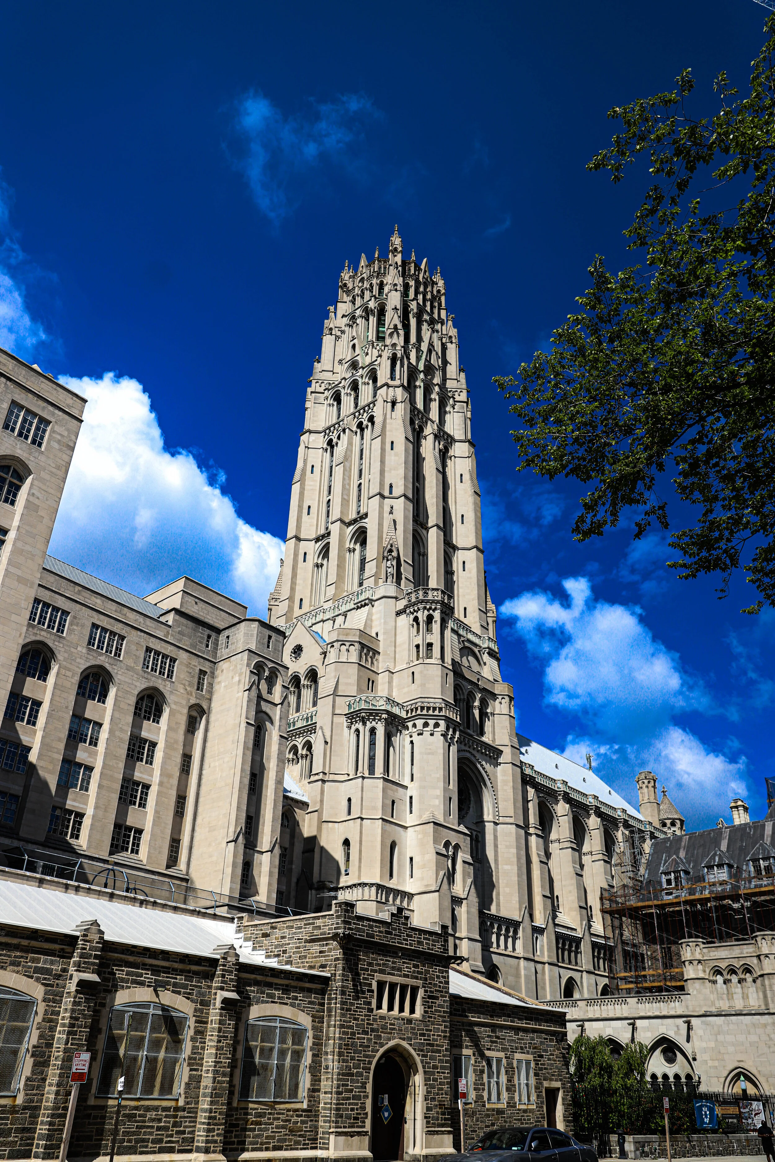 A tall, gothic-style church tower with intricate architecture, situated above stone buildings with barred windows, set against a blue sky with wispy clouds and a leafy tree on the right.