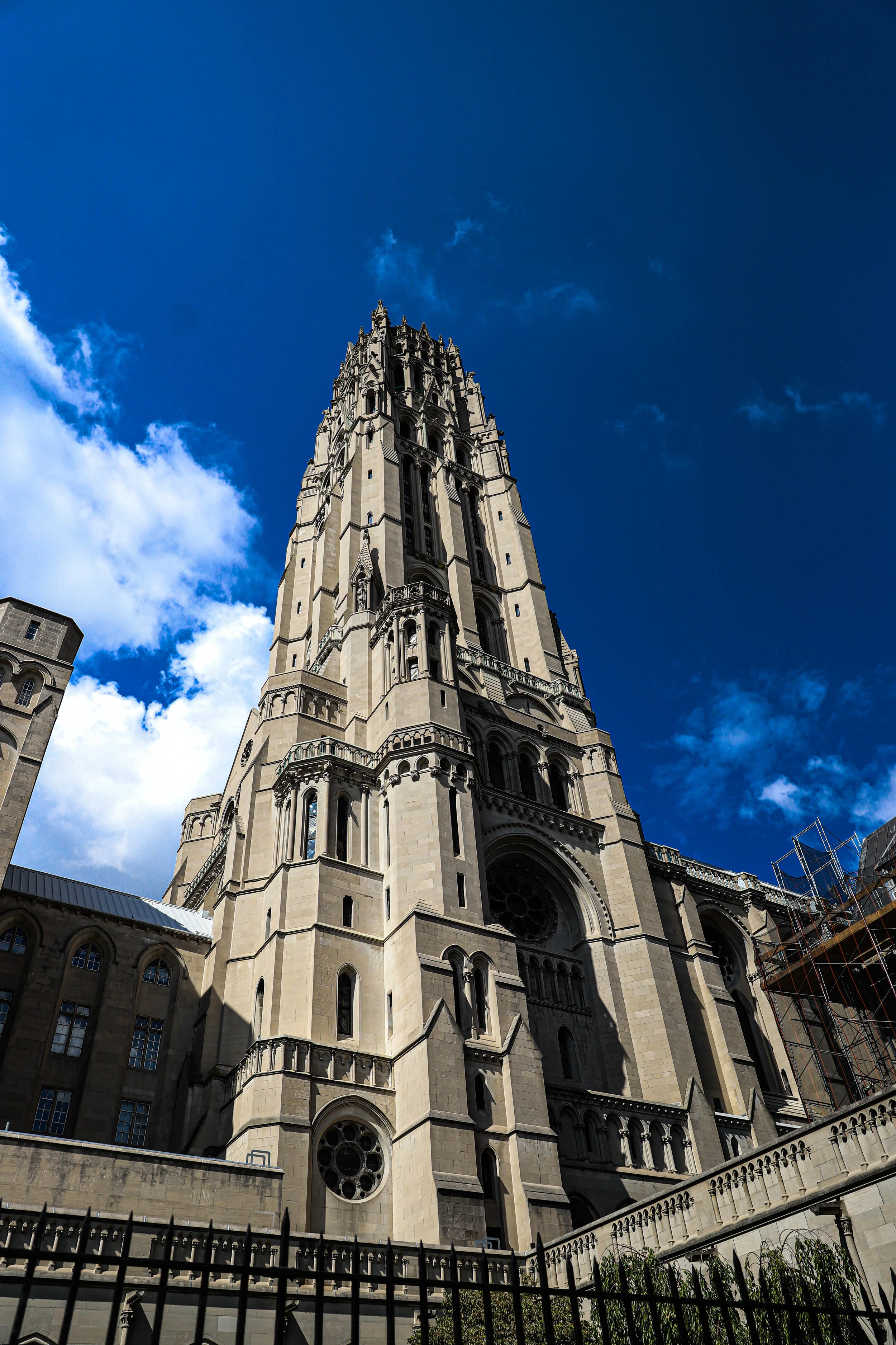 A tall Gothic-style church or cathedral with intricate stonework, pointed arches, large windows, and a spire set against a bright blue sky with some clouds.
