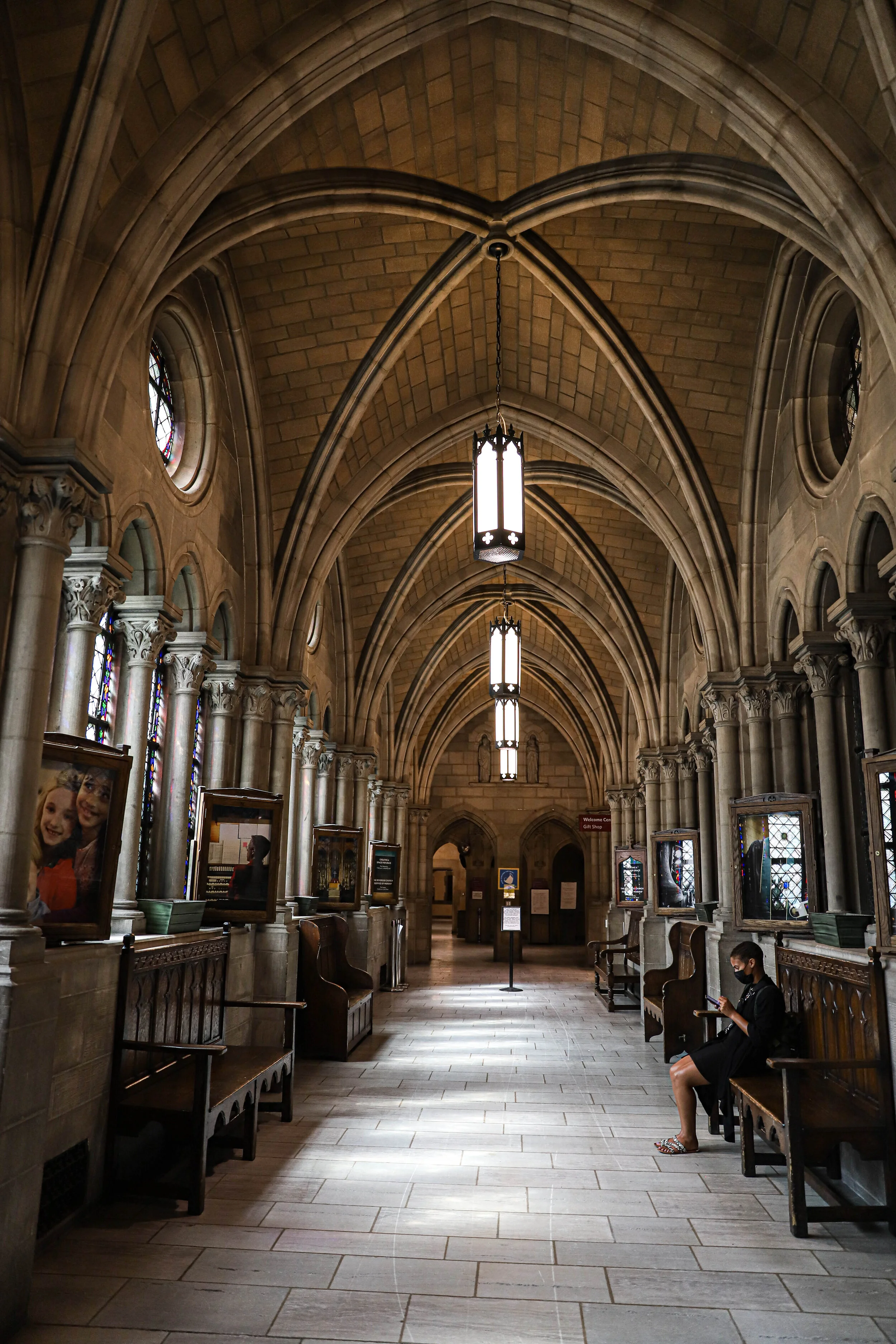 Inside a Gothic-style cathedral with high vaulted ceilings, stained glass windows, and rows of wooden benches. A woman wearing a face mask is sitting on one of the benches, looking at her phone.