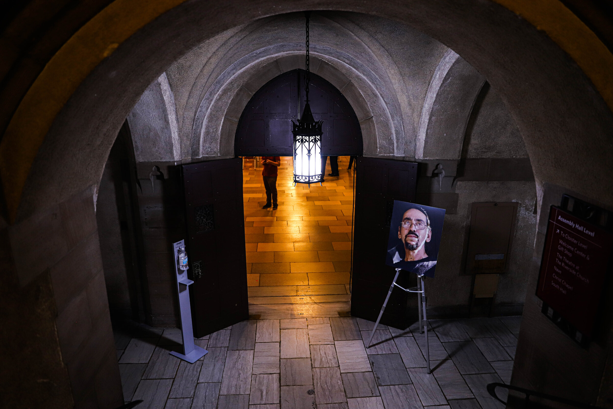 View through a stone archway into a hallway with a hanging lantern, a poster of a man's face on an easel, and people visible in the background.