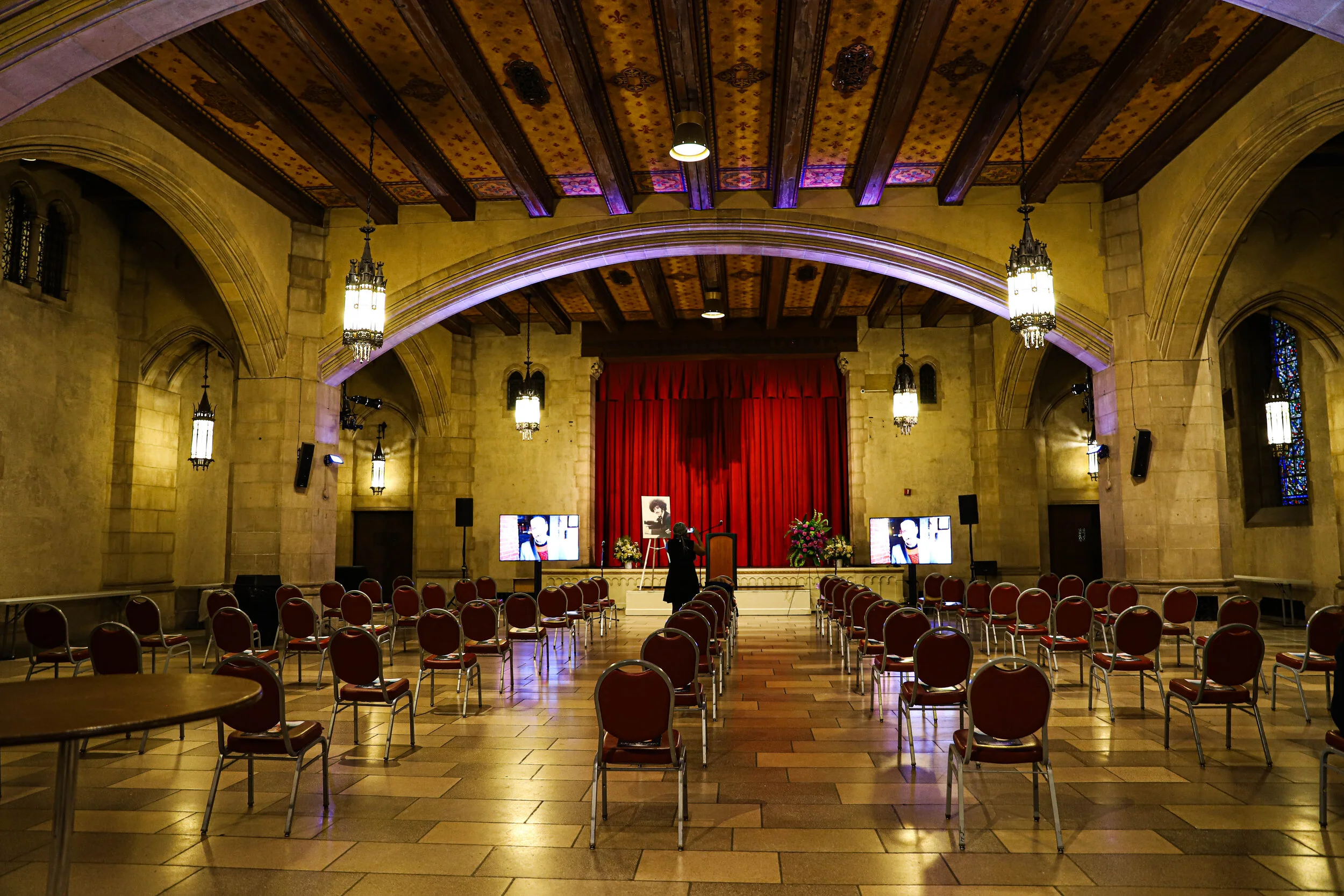 Interior of a large hall with arched ceilings and hanging lantern lights. Red curtains on stage, with chairs arranged for an event. Multiple screens display a person's image toward the front of the room.