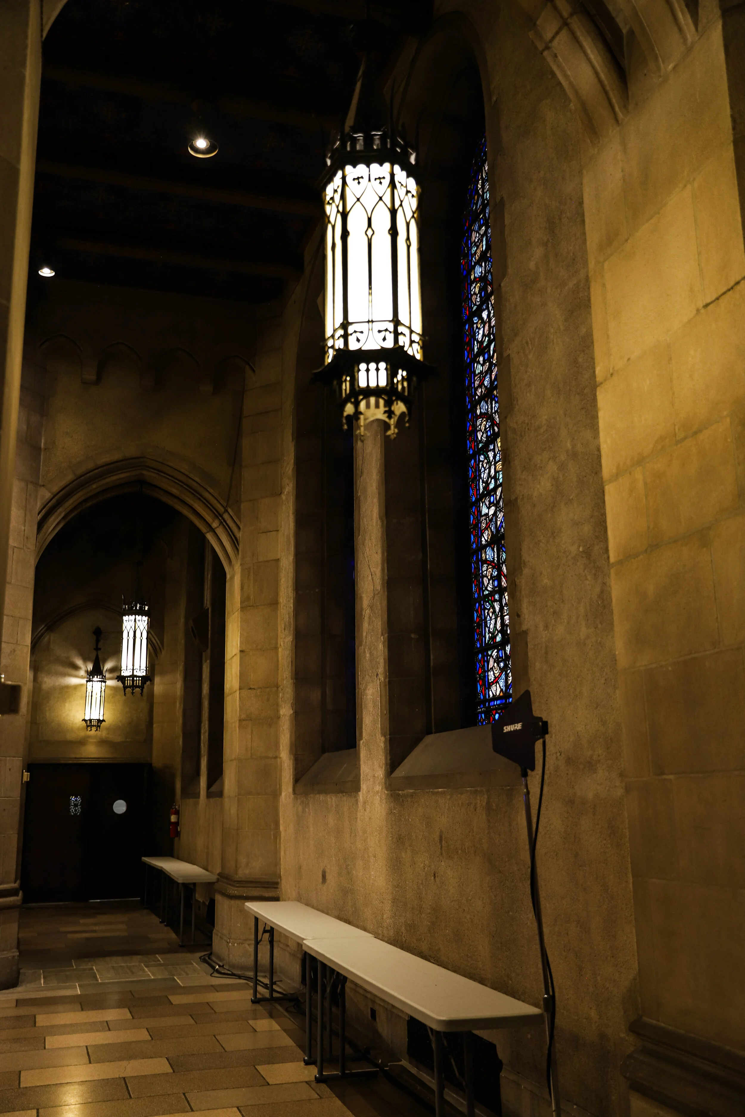 Interior of a church or cathedral with stone walls, stained glass windows, hanging lantern-style lights, and a wooden floor. There are white benches along the wall and a microphone on a stand.