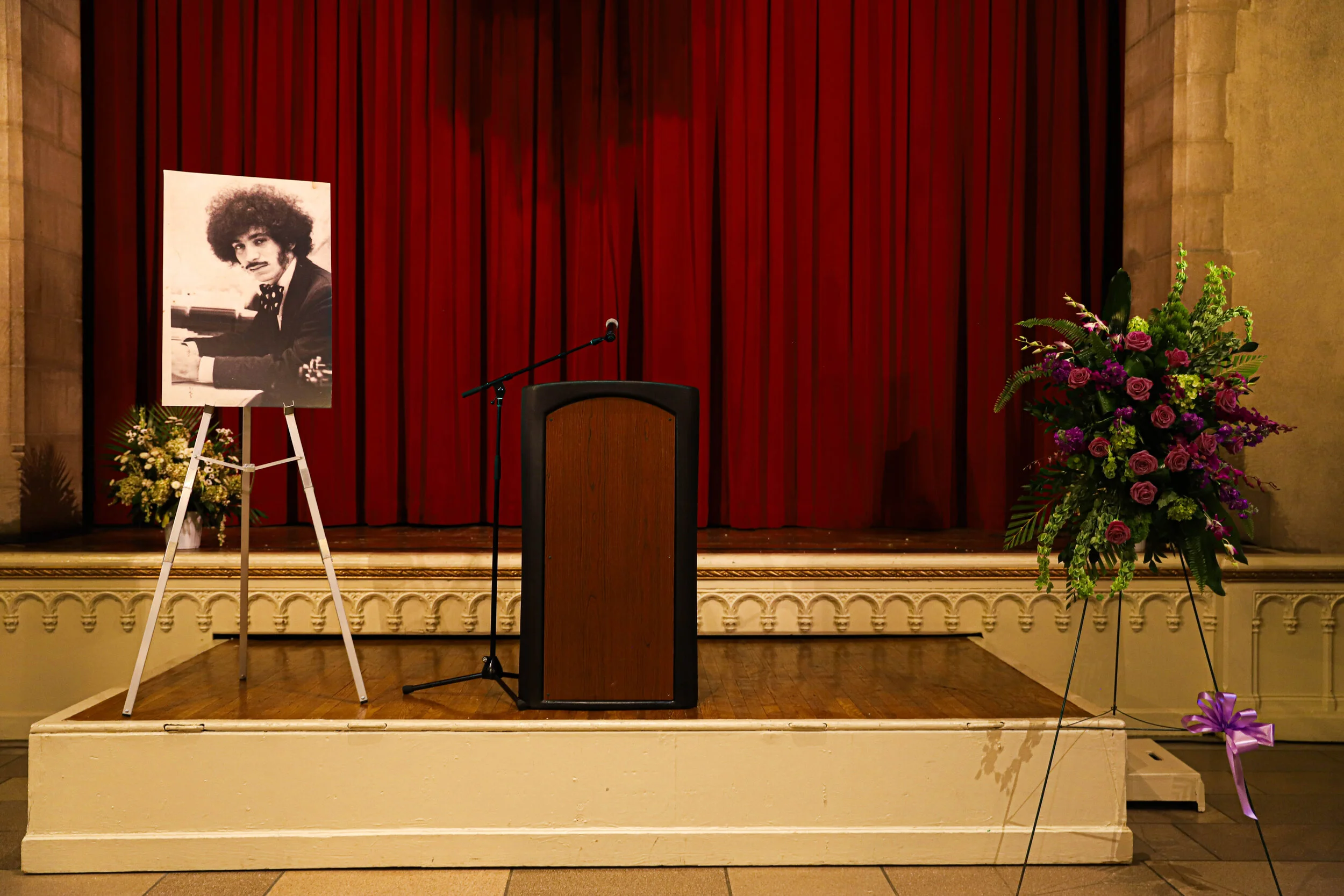 Empty stage with a red curtain, a wooden podium, a microphone, a large floral arrangement, and a portrait of a man playing the piano.