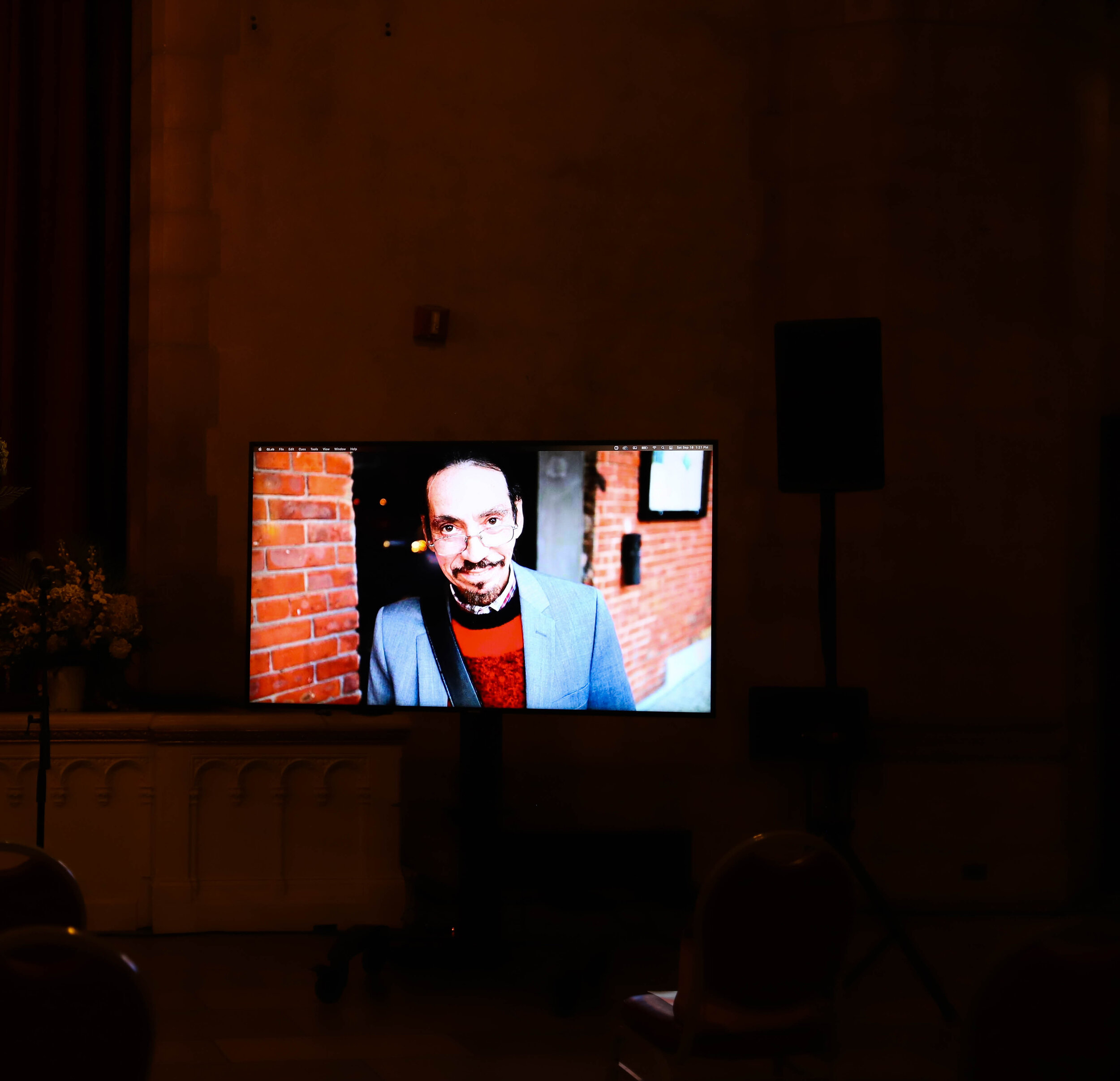 Dark room with a television screen displaying a man wearing glasses and a suit, with a brick wall background.