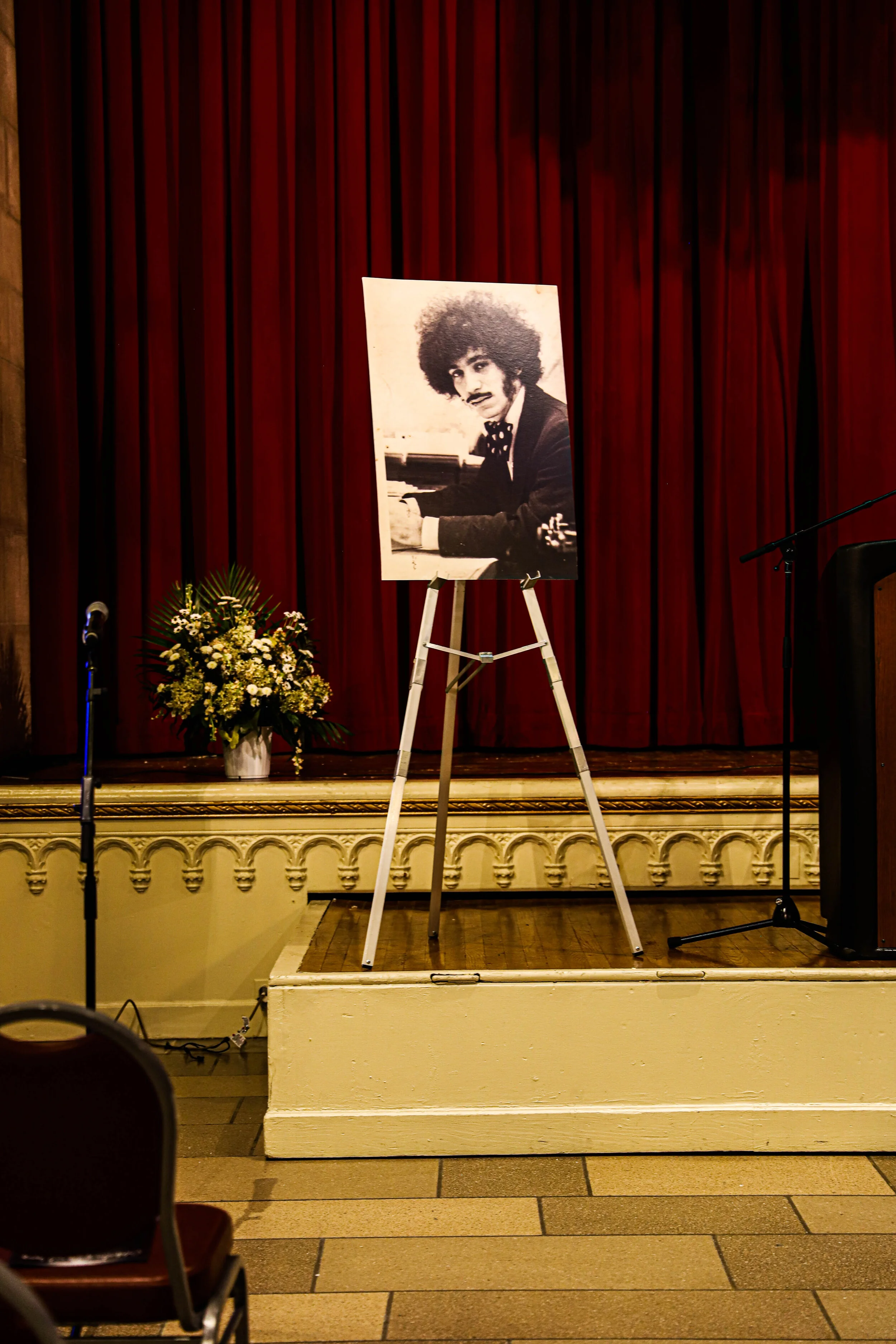 A stage with red curtain backdrop featuring a large black-and-white portrait of a man with curly hair and mustache, sitting at a piano. There is a flower arrangement to the left and microphones on stage.
